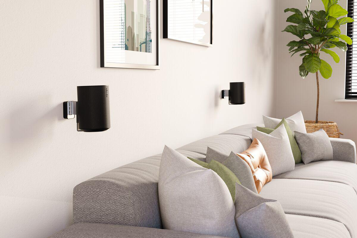 Wohnzimmer mit grauer Couch, Kissen, zwei Wandlautsprechern, Bildern und Zimmerpflanze.