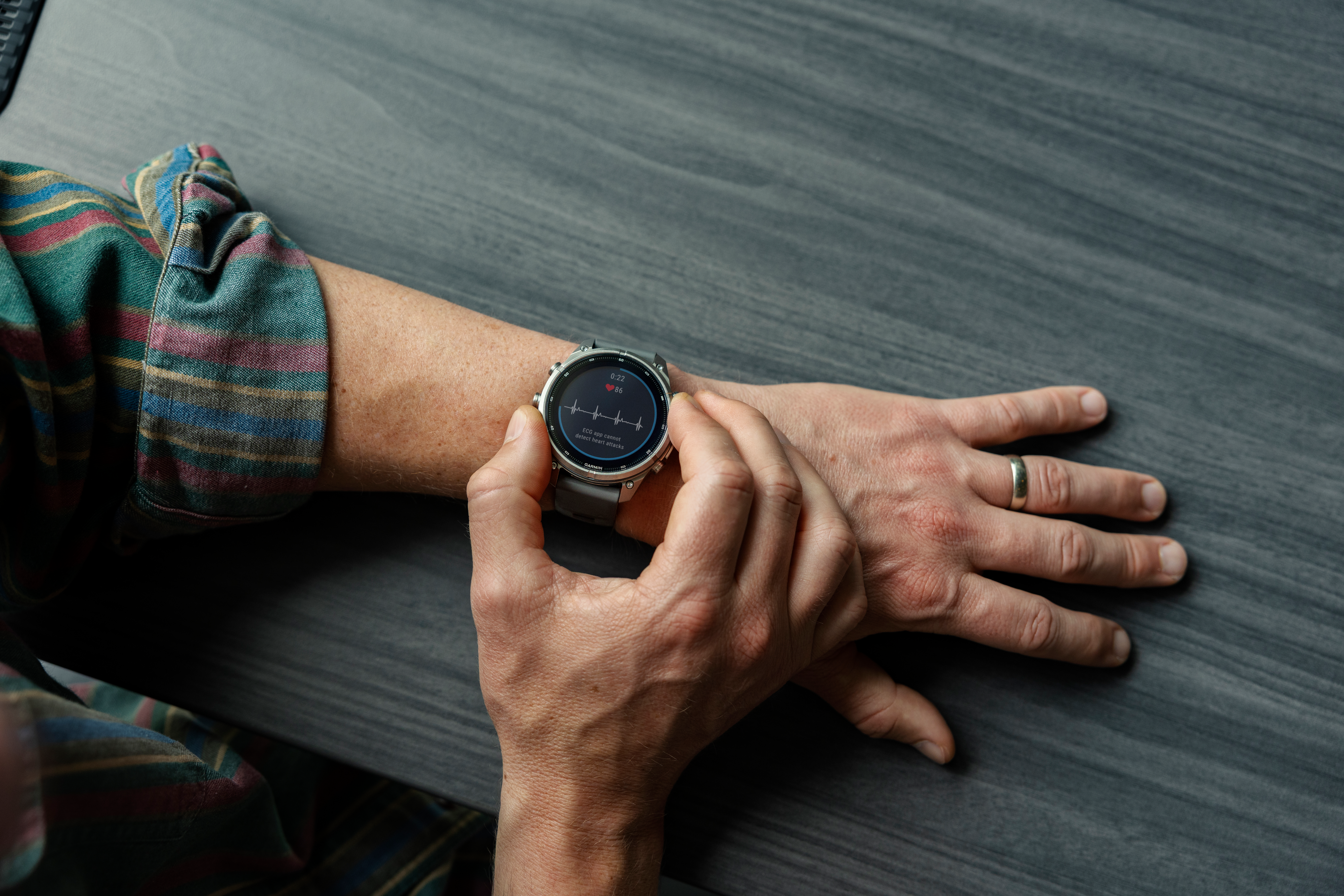 Eine Person berührt mit den Fingern den Bildschirm einer Smartwatch mit EKG-Grafik auf einem dunklen Holztisch.