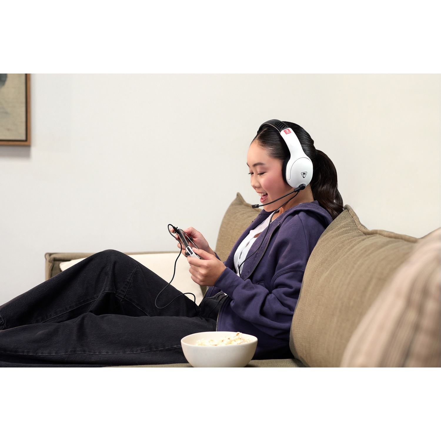 A young woman wearing a headset sits on a sofa, playing a handheld device, with a bowl of popcorn in front of her.