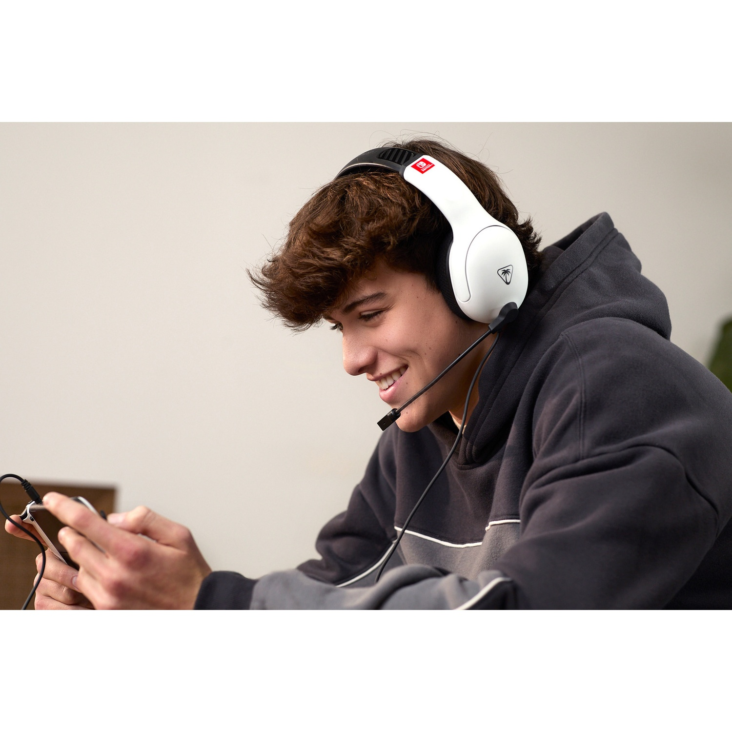 Young boy with white headset and black hoodie smiling while playing on a small mobile device.