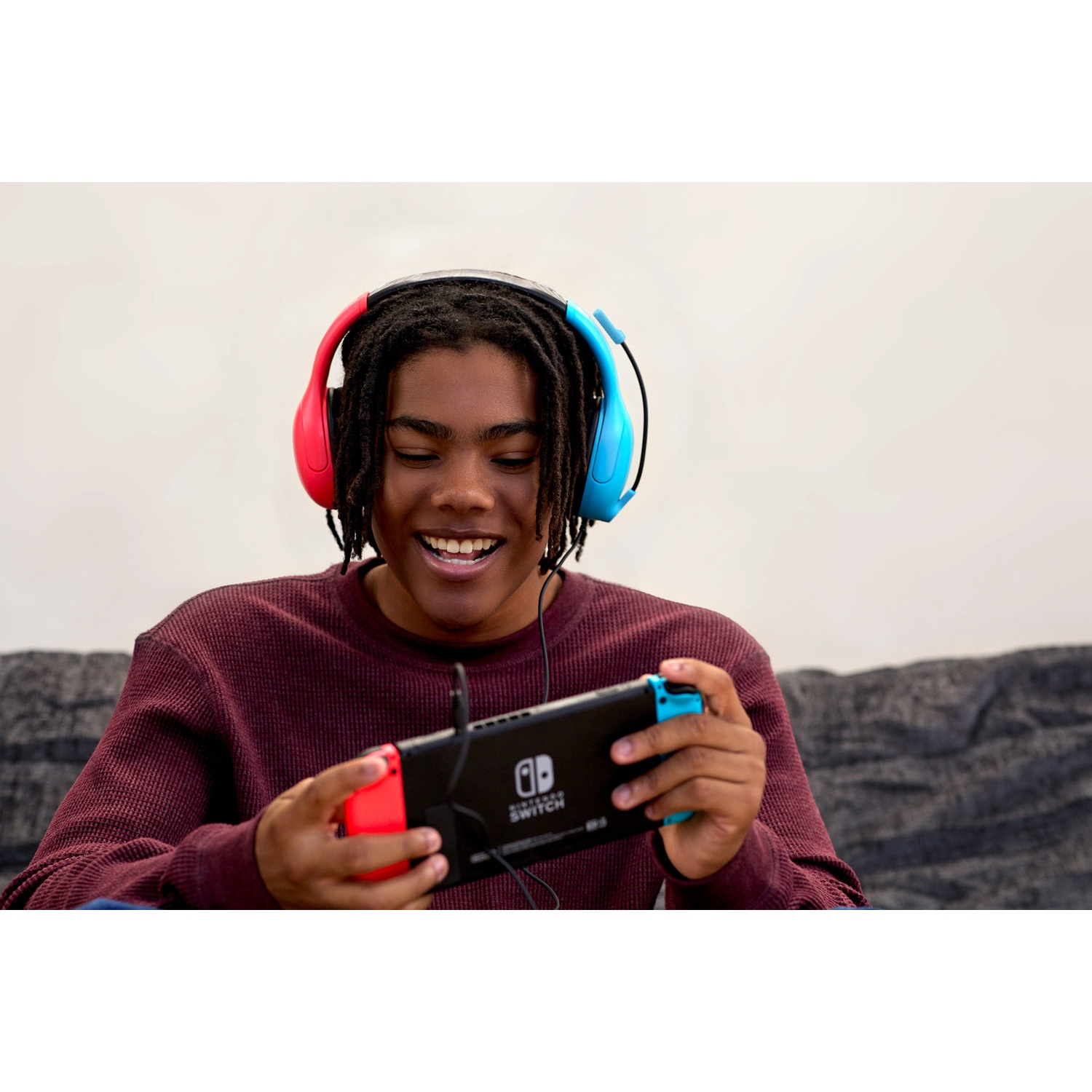 A smiling young man with dreadlocks and red-and-blue headphones plays a handheld video game on a sofa.