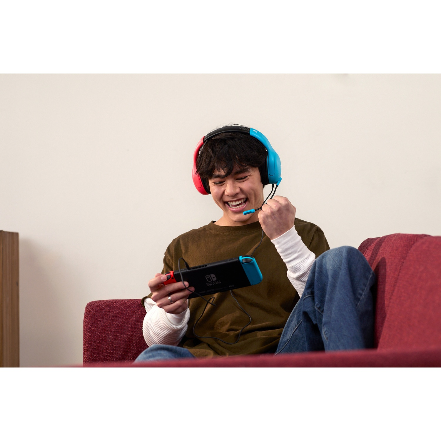 A young man sits on a red sofa, cheering while playing on a handheld console.