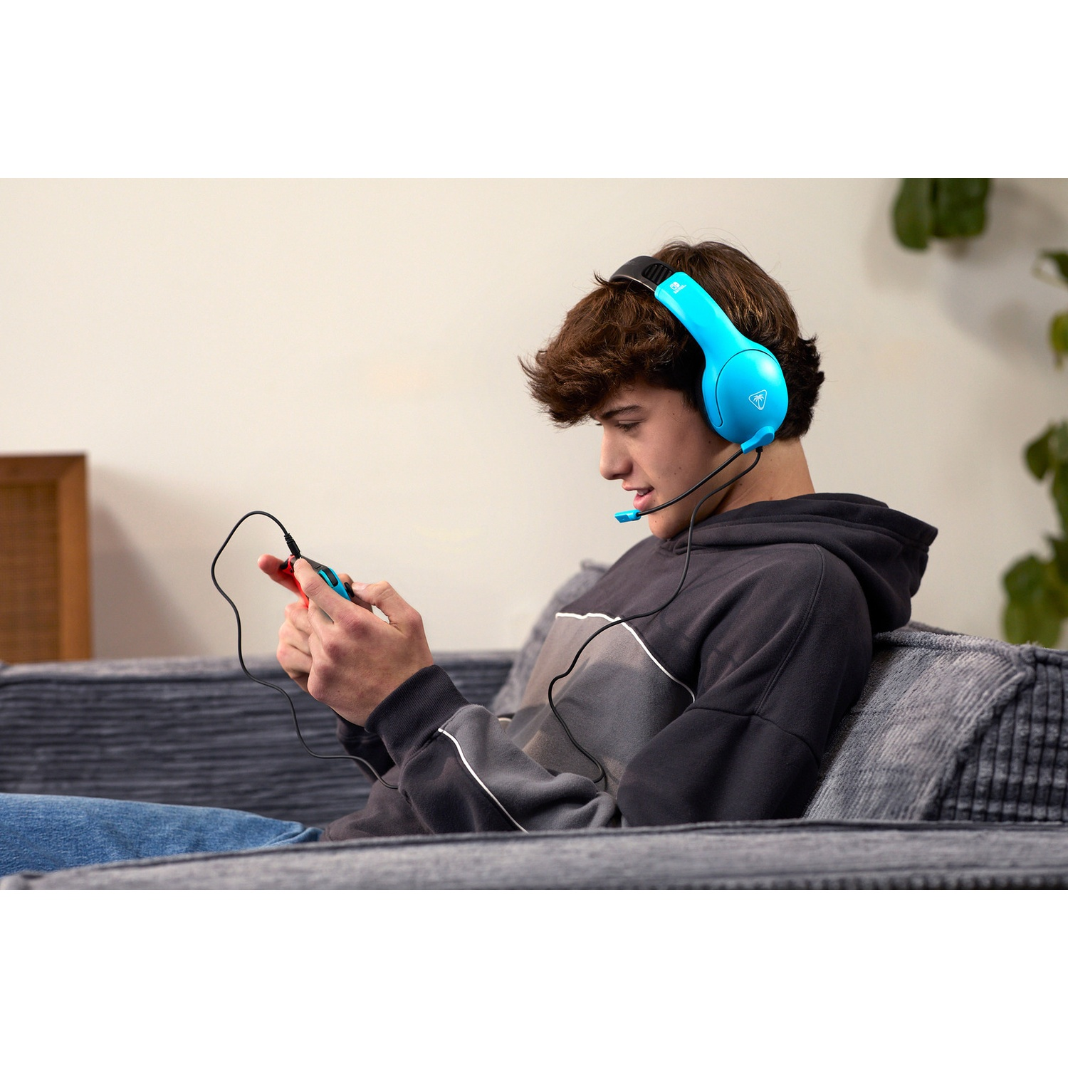 A young man sitting on a gray couch, wearing bright blue headphones with a mic, playing a handheld game console.