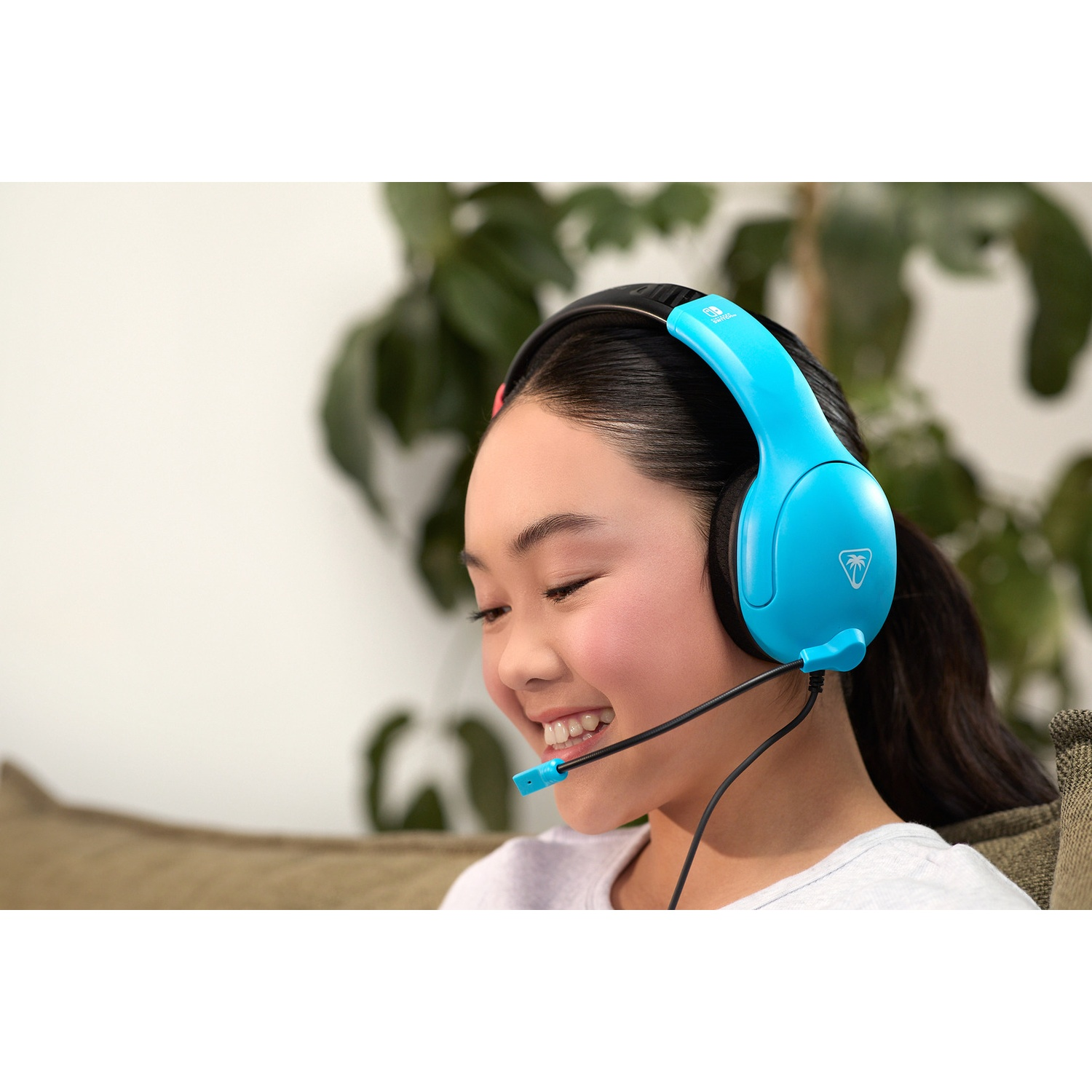 A young girl with black hair smiles, wearing blue gaming headphones with a microphone in front of a plant.