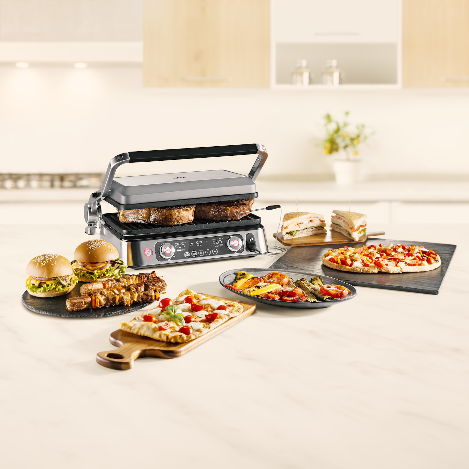 Una griglia elettrica cuoce bistecche con hamburger, pizza, panini e verdure su un bancone da cucina.