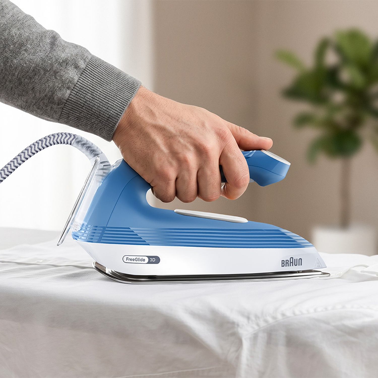 Une main en pull gris tient un fer à repasser Braun bleu et blanc sur du linge blanc.