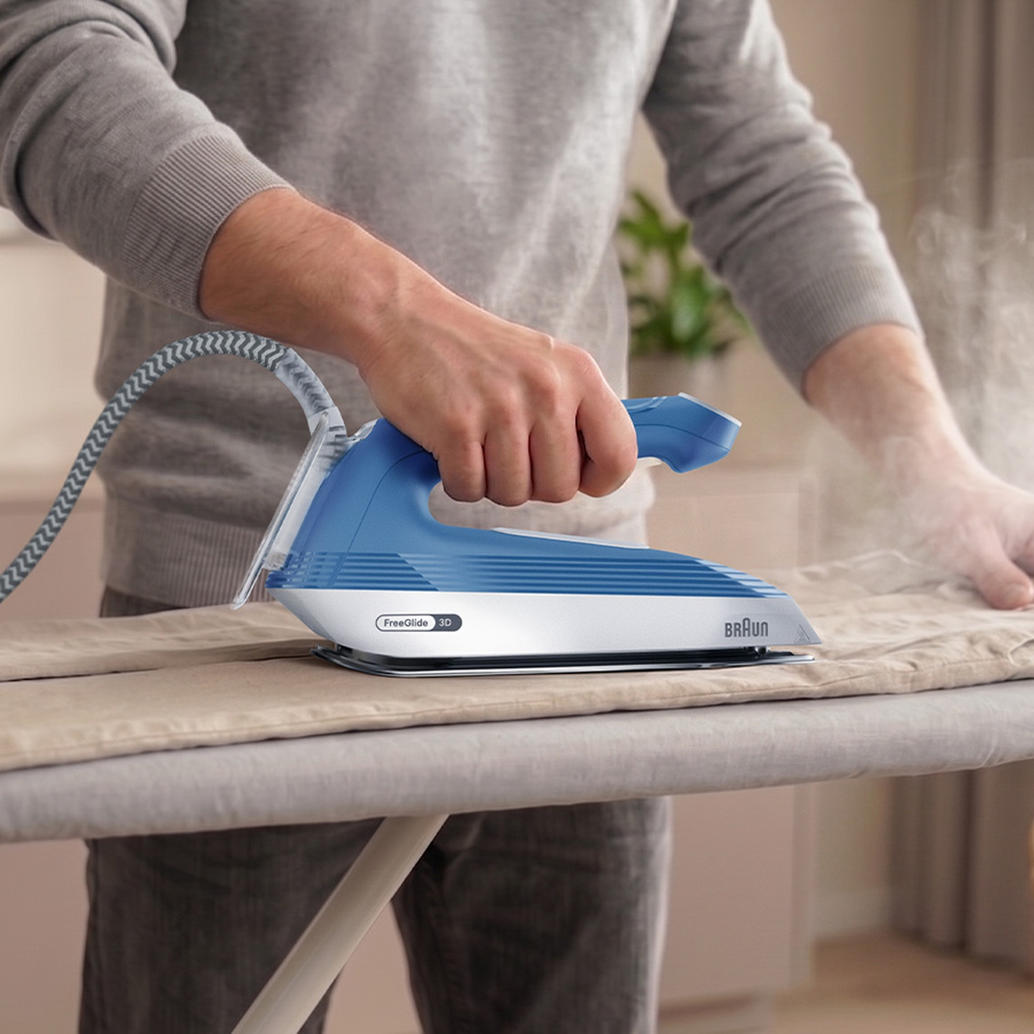 Une personne en pull gris utilise un fer à repasser Braun bleu et blanc sur une table.