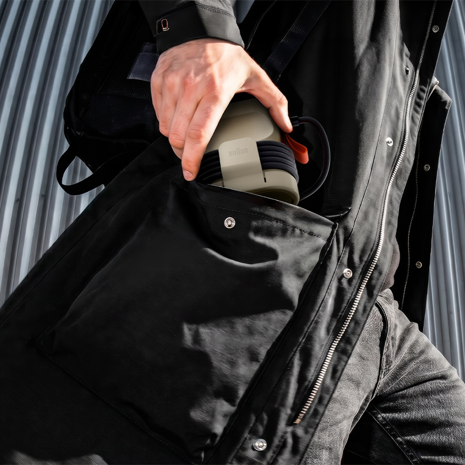 A person in a black jacket places a Braun portable speaker into a black backpack pocket against a corrugated wall.
