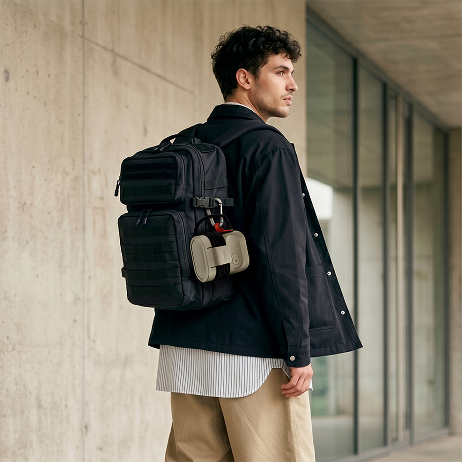 A young man with a dark jacket and black backpack stands before a concrete wall, looking to the side.