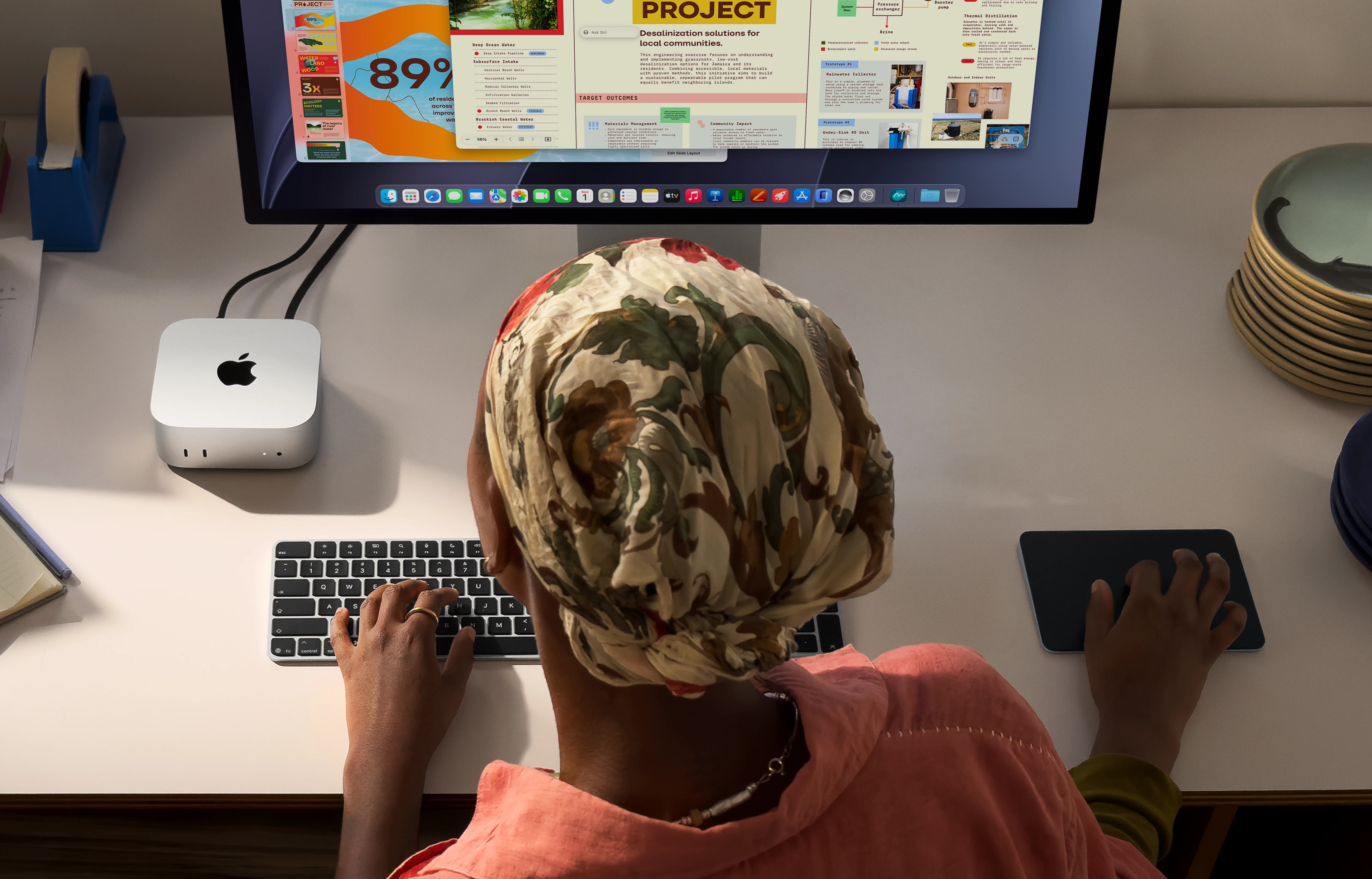 Mintás fejkendős személy ír az asztalnál egy nagy monitor és Mac Mini előtt.