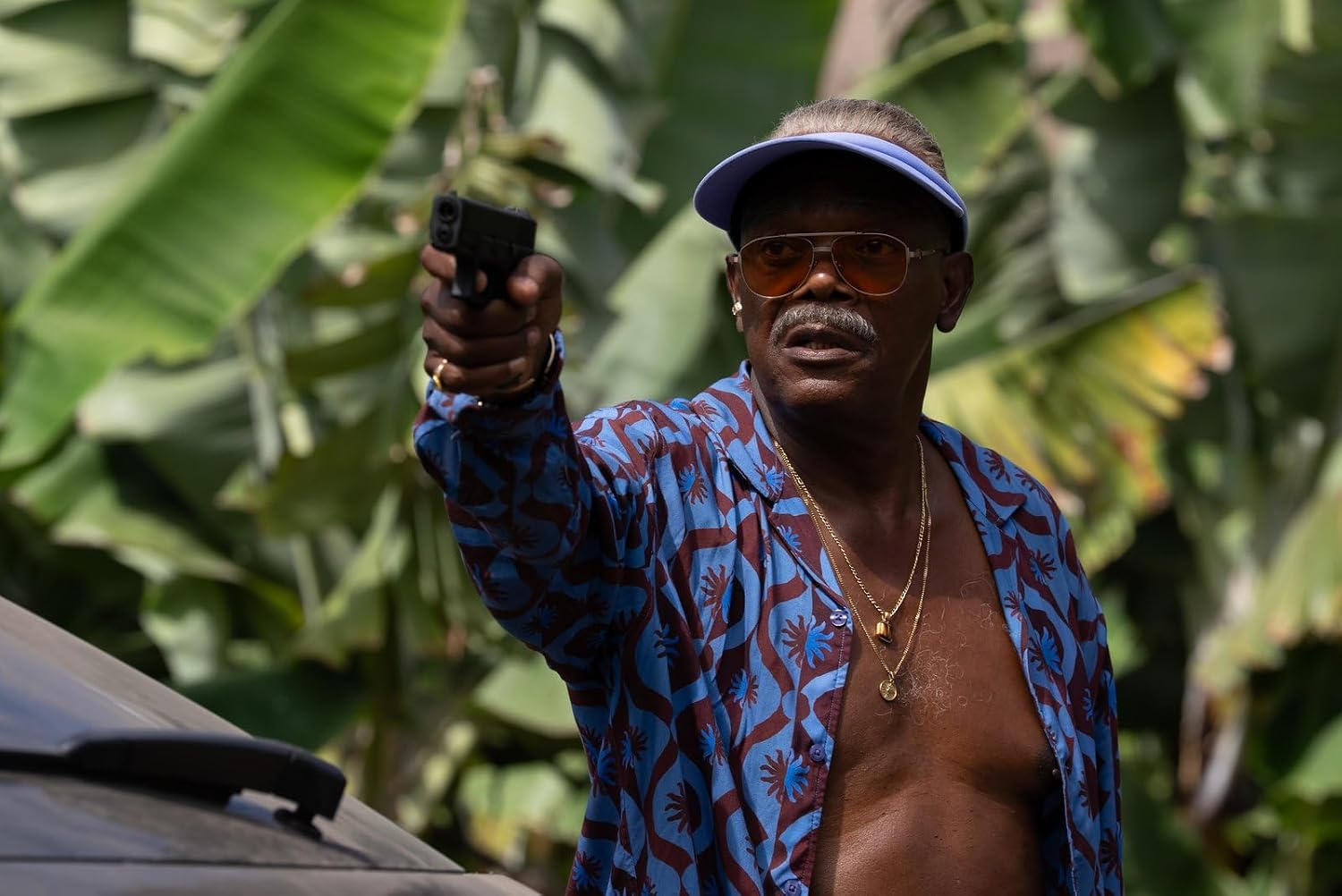 Samuel L. Jackson zielt mit einer Pistole, trägt ein blaues Hemd und eine Sonnenbrille vor grünen Blättern.