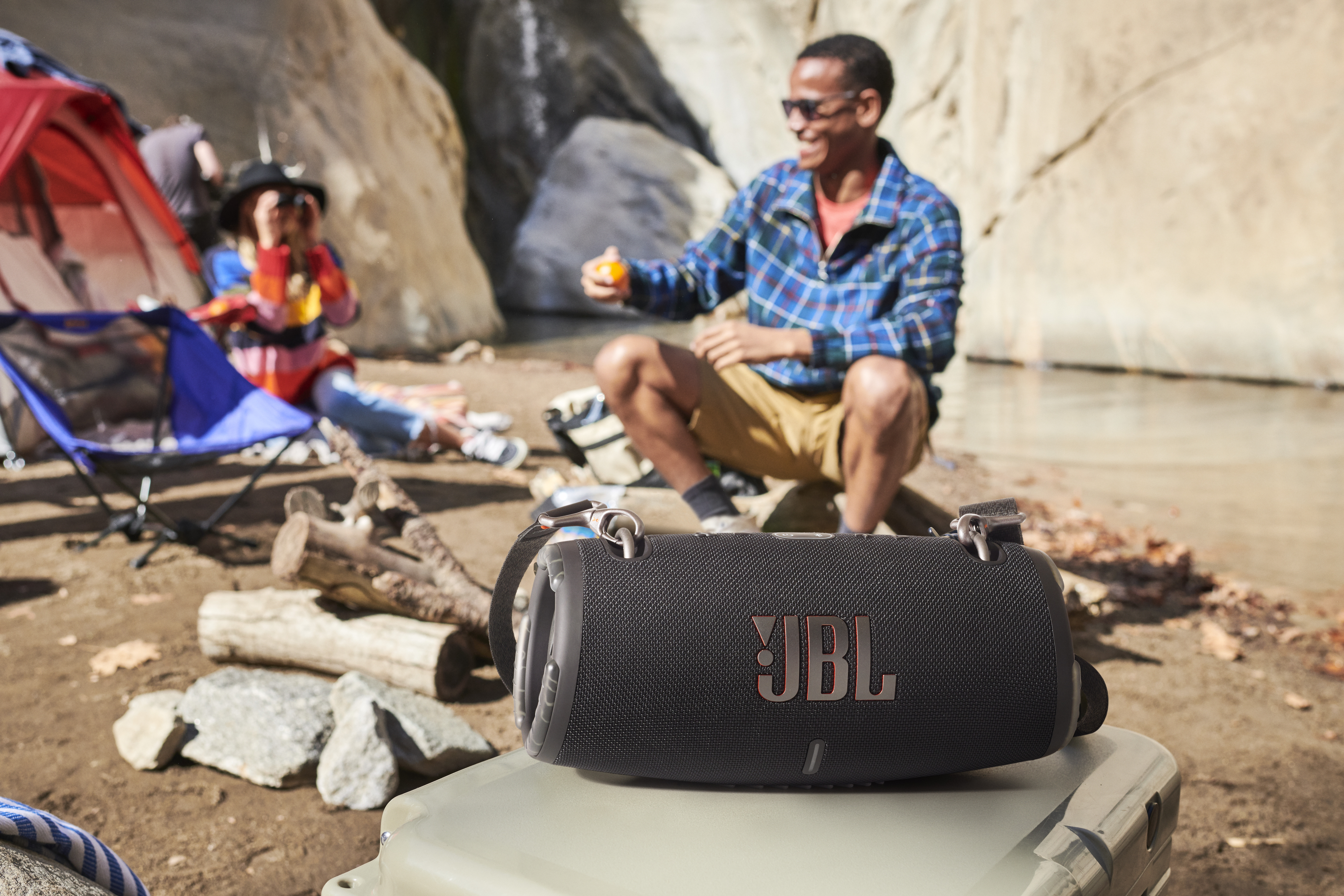 Czarny głośnik bluetooth JBL spoczywa na lodówce turystycznej na pierwszym planie, a w tle ludzie koczują przy namiocie.