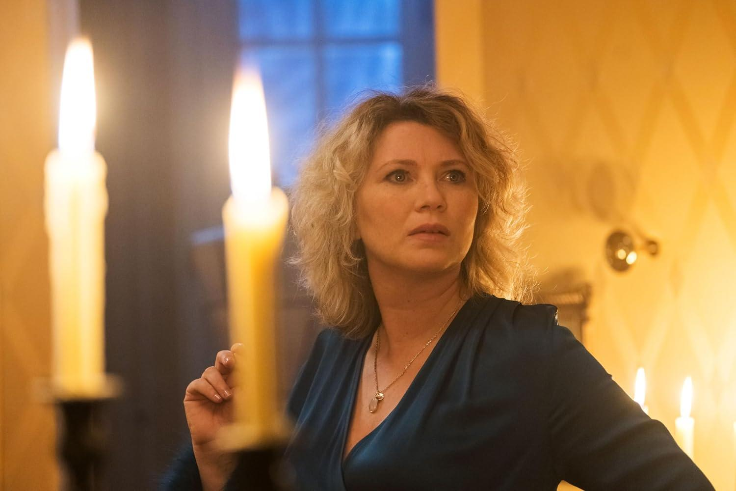 Eine blonde Frau in einem blauen Kleid steht in einem warm beleuchteten Raum mit Kerzen im Vorder- und Hintergrund.