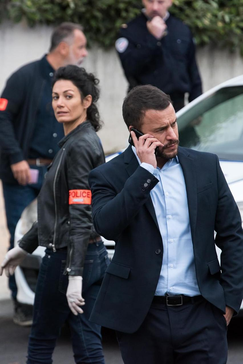 Ein Mann im Anzug telefoniert vor einem Polizeiwagen, während andere Beamte im Hintergrund wachen.