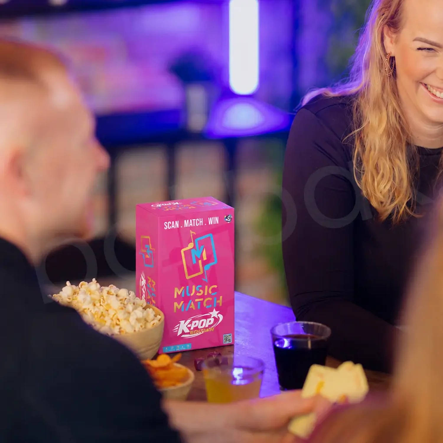Friends play a K-Pop music game at a table with popcorn and drinks in a room with blue lighting.