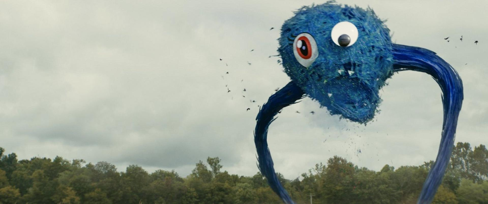 Eine große, blaue Kreatur mit großen Augen schwebt über einem Wald unter bewölktem Himmel.