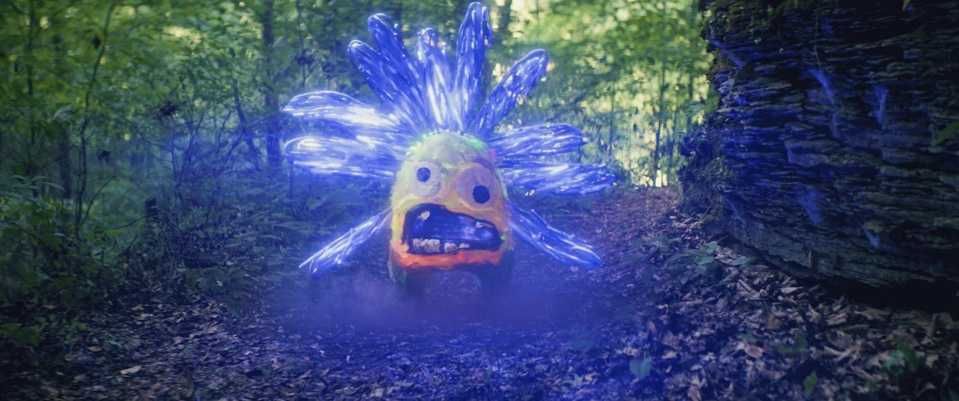 Ein skurriles, pelziges Wesen mit blauen Kristallblüten steht auf einem Waldweg, umgeben von Bäumen und Nebel.