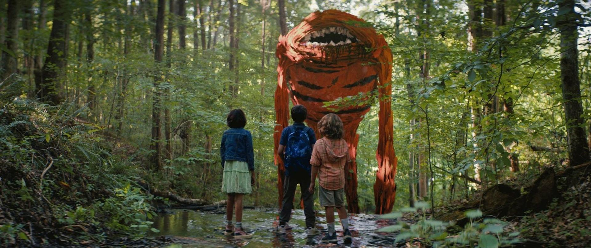 Drei Kinder stehen im Wald und blicken auf ein großes orangefarbenes Monster mit offenem Maul.