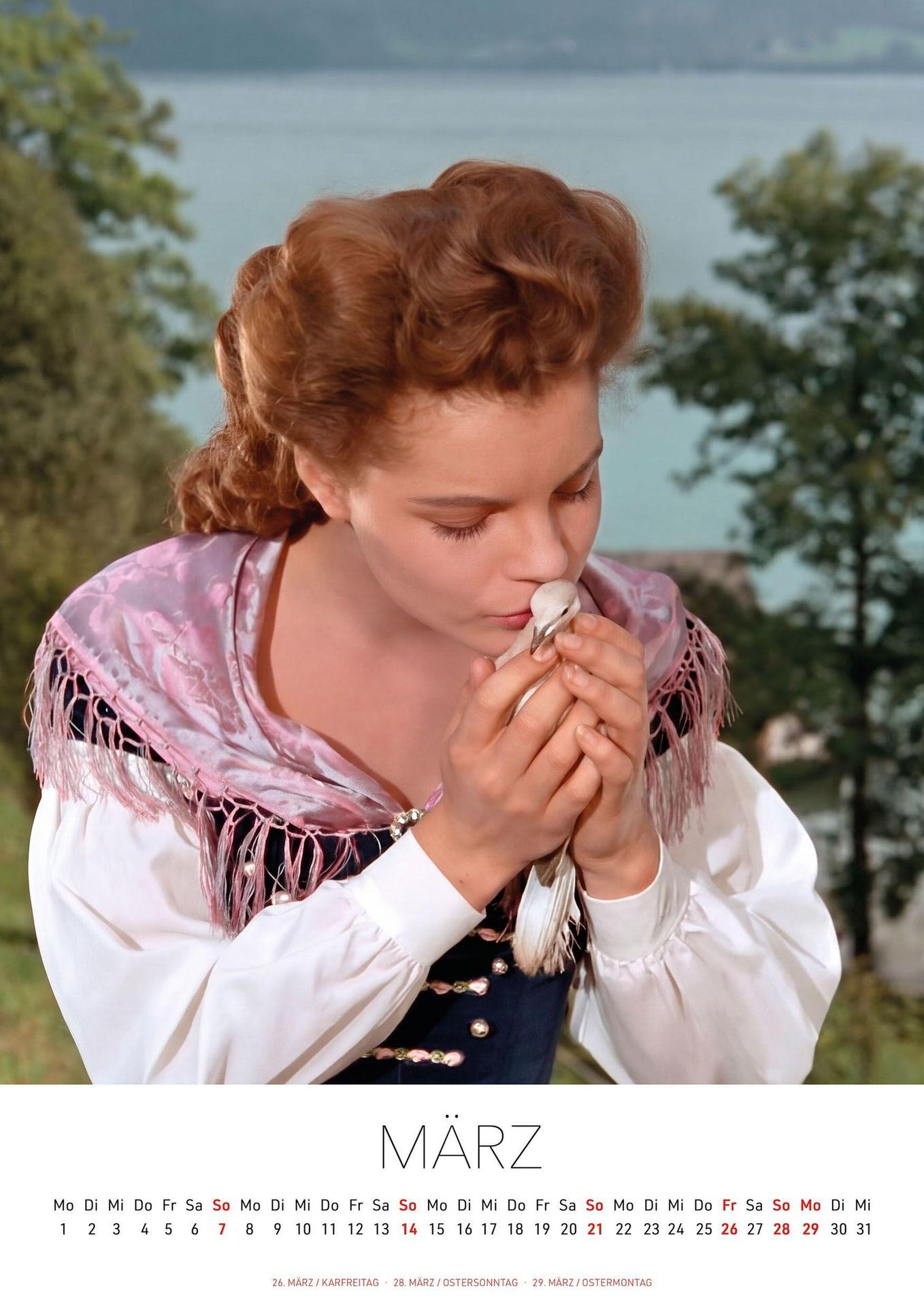 Eine Frau in Tracht küsst zärtlich einen kleinen Vogel vor einem See.