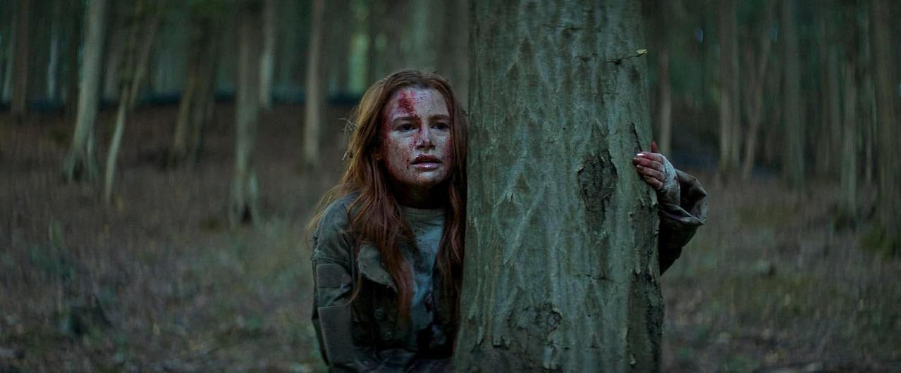Eine blutverschmierte junge Frau mit roten Haaren versteckt sich verängstigt hinter einem Baum in einem dunklen Wald.