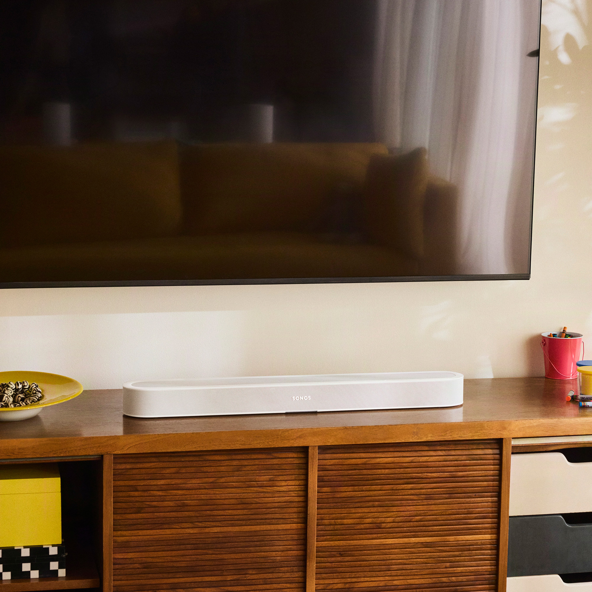Una barra de sonido blanca Sonos descansa sobre un mueble de madera debajo de un televisor.