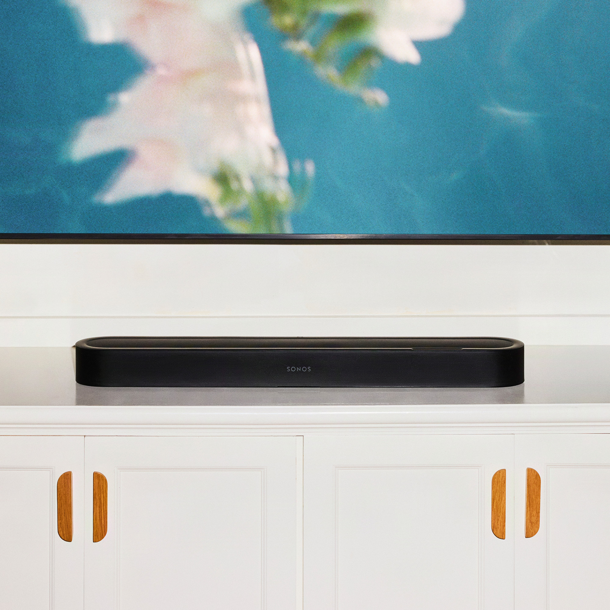Una barra de sonido Sonos negra se encuentra en una consola blanca frente a una pantalla azul.