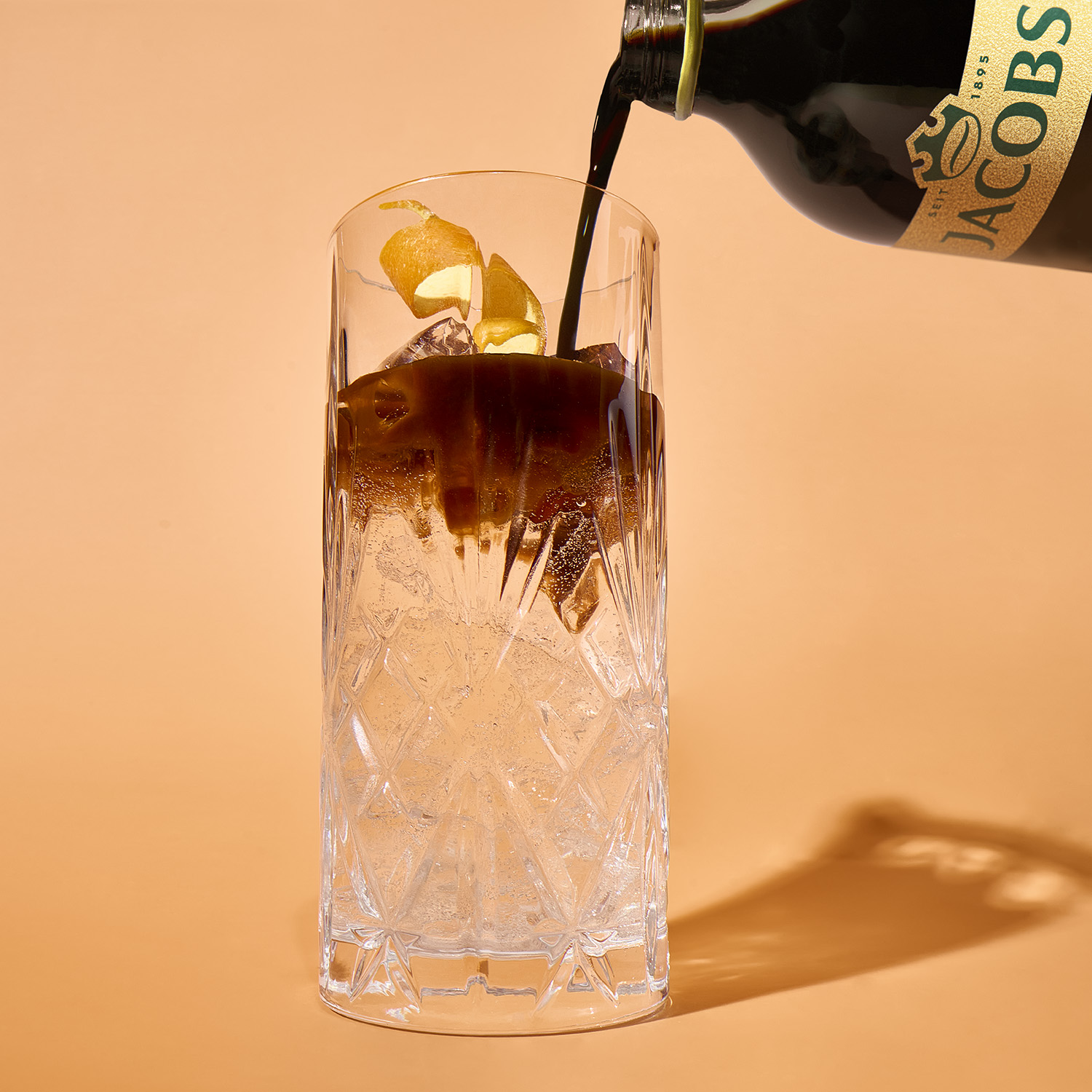 Dunkle Flüssigkeit wird aus einer Flasche in ein hohes Schliffglas mit Eis und einer Zitronenschale auf beige gegossen.