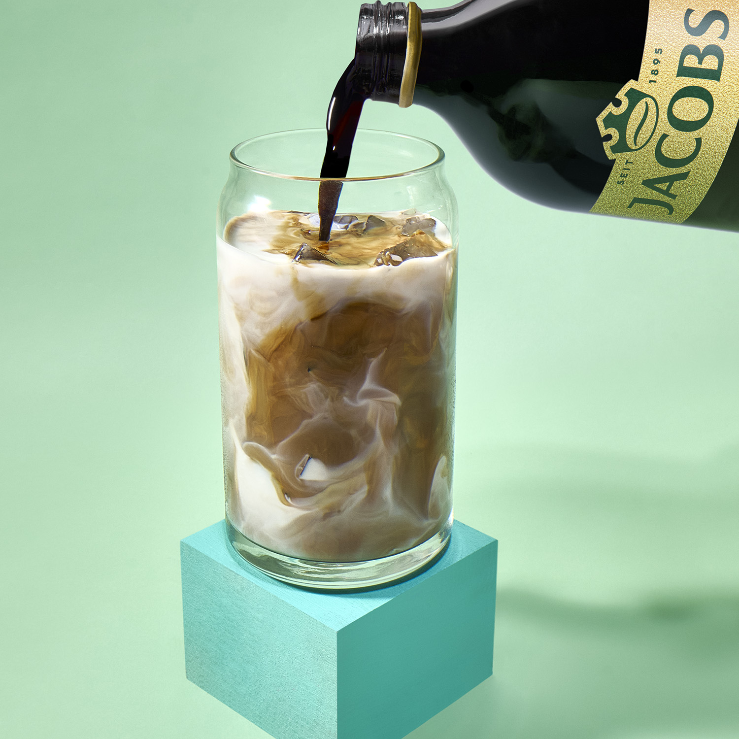 Jacobs-Kaffee wird aus einer Flasche in ein Glas mit Eis und Milch auf einem türkisen Sockel gegossen.