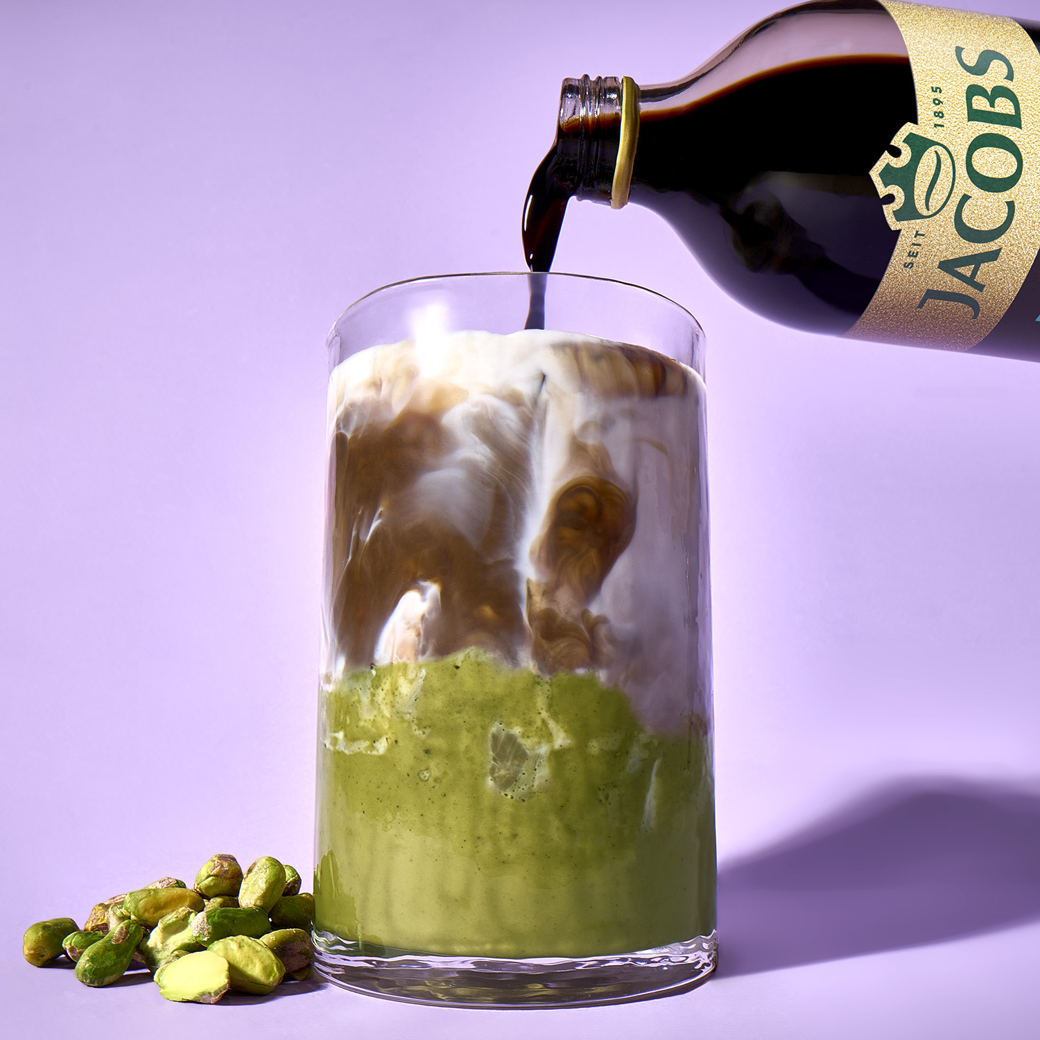 Jacobs-Kaffee wird in ein Glas mit Milch und Pistazienpaste gegossen, daneben liegen Pistazien.