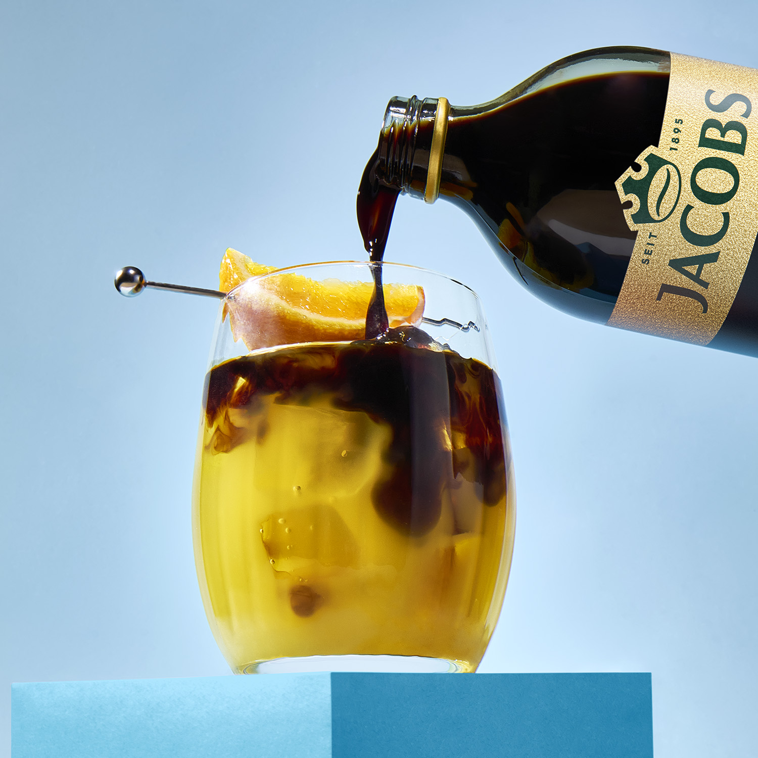 Jacobs-Kaffee wird aus einer Flasche in ein Glas mit Eis und Orangenscheibe auf blauem Grund gegossen.