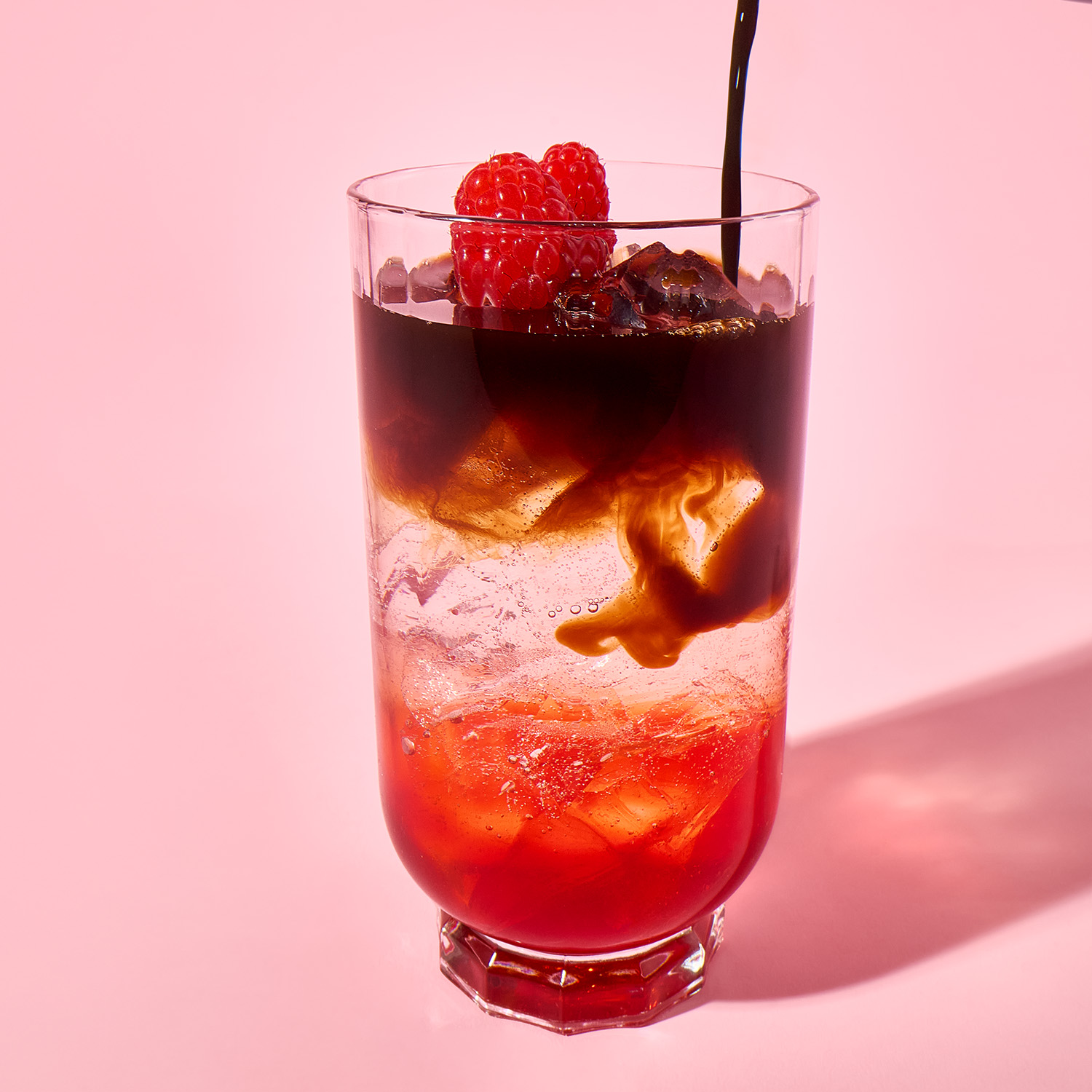 Dunkle Flüssigkeit fließt in ein hohes Glas mit Eis und rotem Sirup, garniert mit Himbeeren, vor rosa Hintergrund.