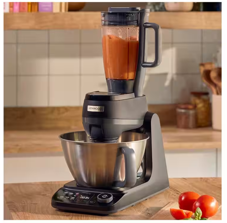 Un robot da cucina grigio scuro con ciotola in metallo e frullatore è su un bancone in legno con pomodori.