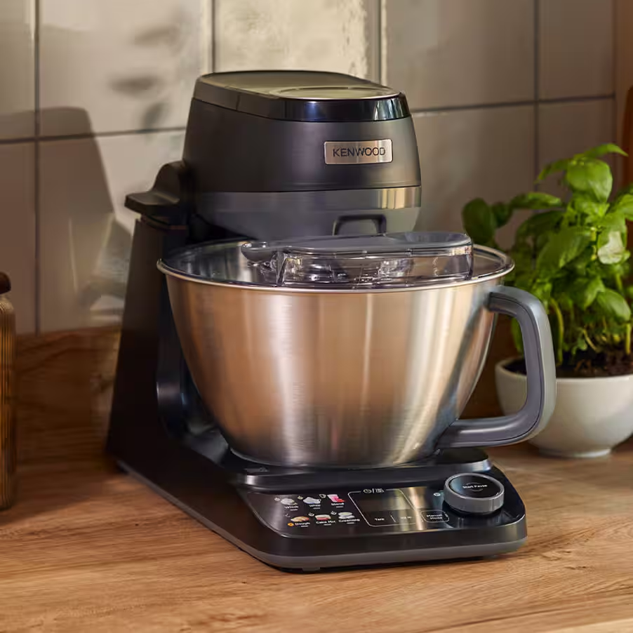 Una planetaria Kenwood nera con ciotola in acciaio inox si trova su un bancone di legno accanto a una pianta in vaso.