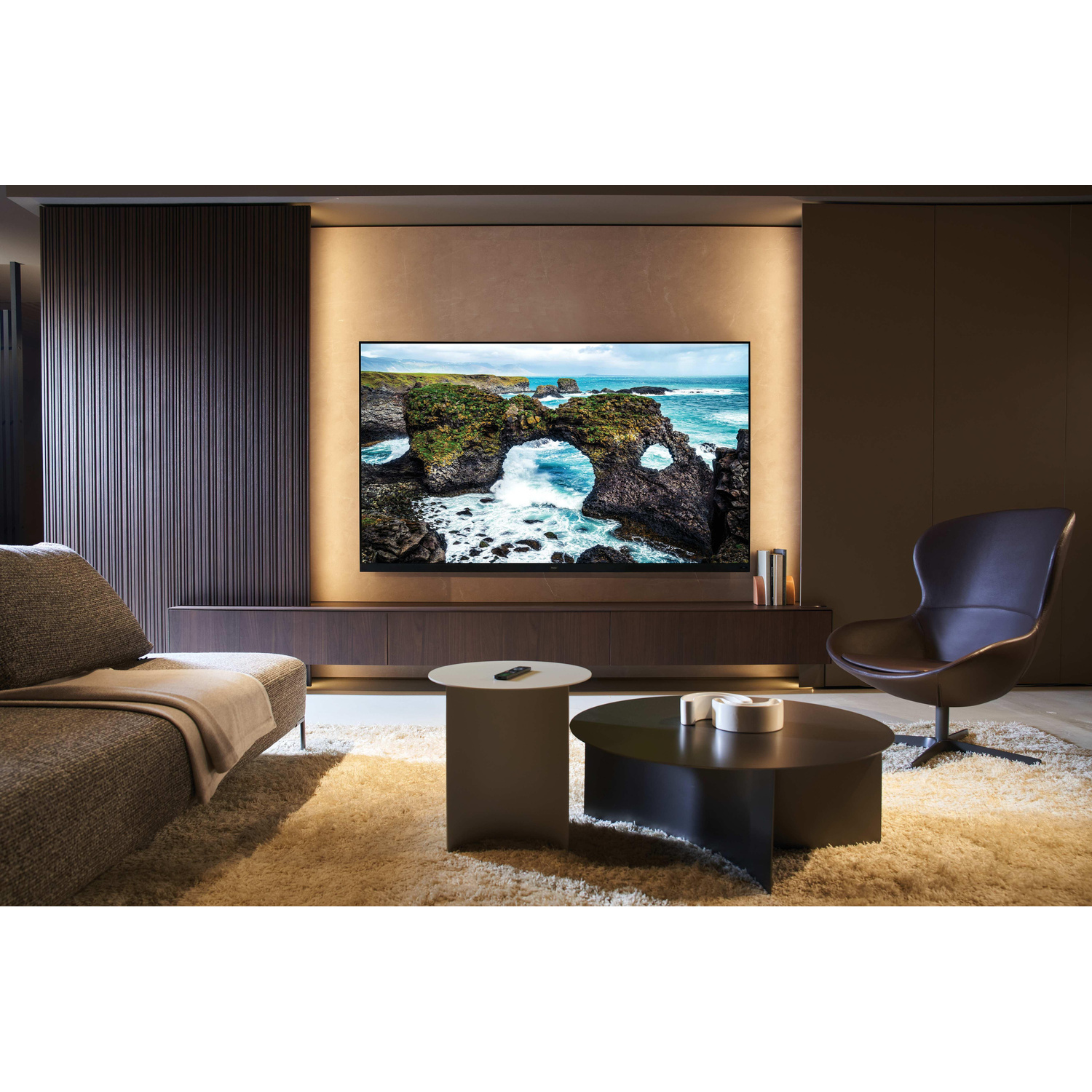 Soggiorno con una grande TV che mostra un paesaggio costiero, due tavolini da caffè e una poltrona marrone.