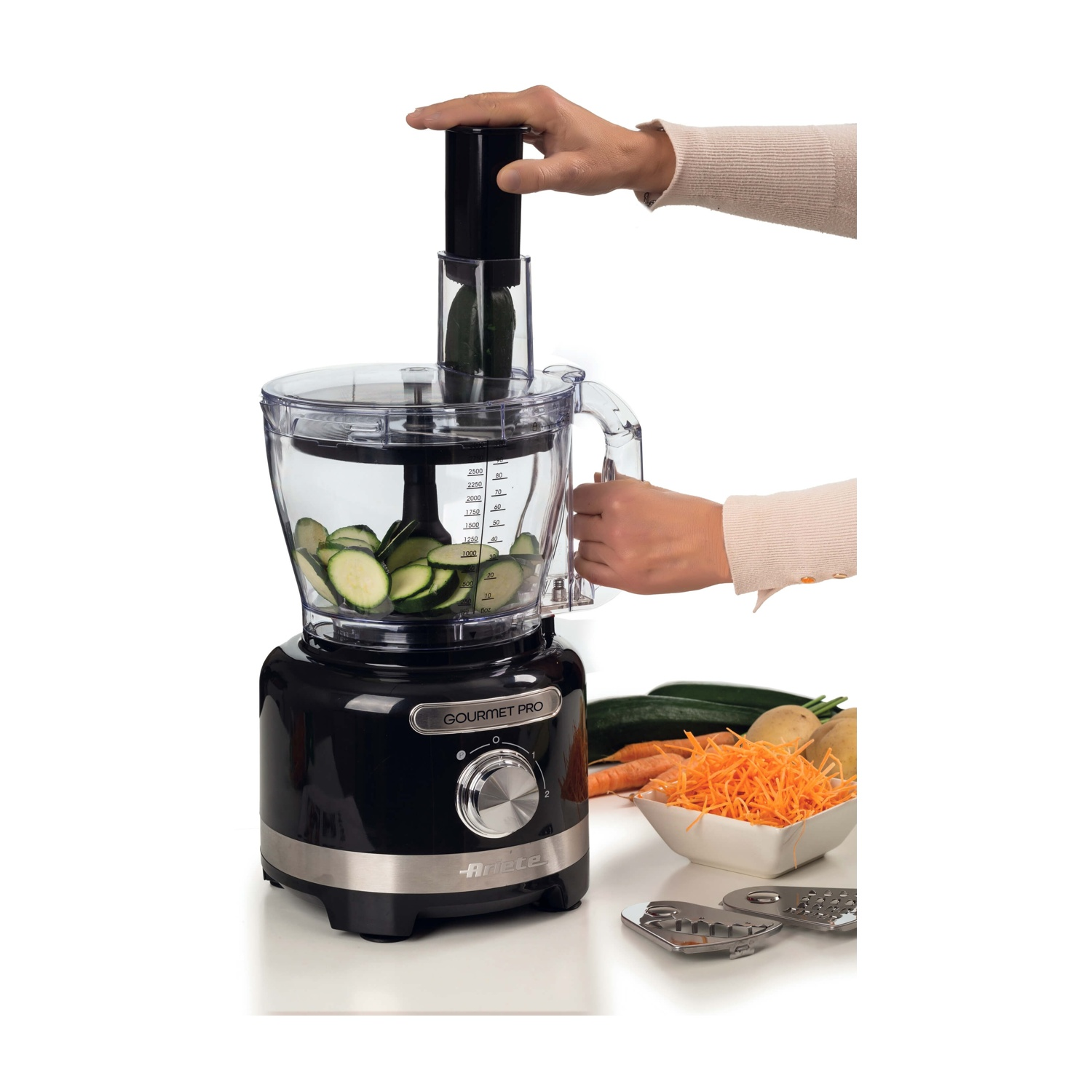 Una mano aziona un robot da cucina nero con fette di cetriolo all'interno, verdure vicine.