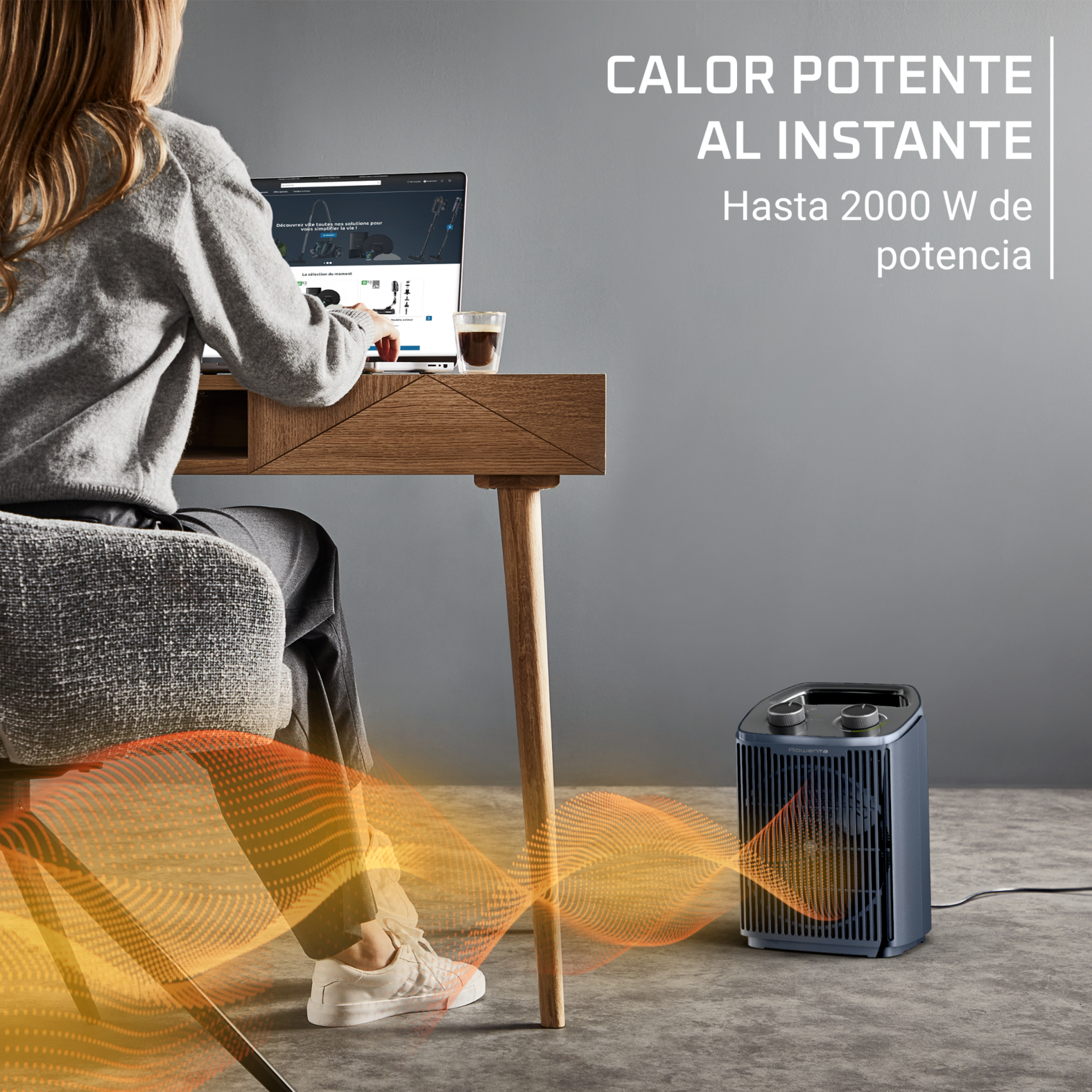 Una mujer trabaja en un portátil en un escritorio de madera mientras un calefactor azul emite aire cálido.