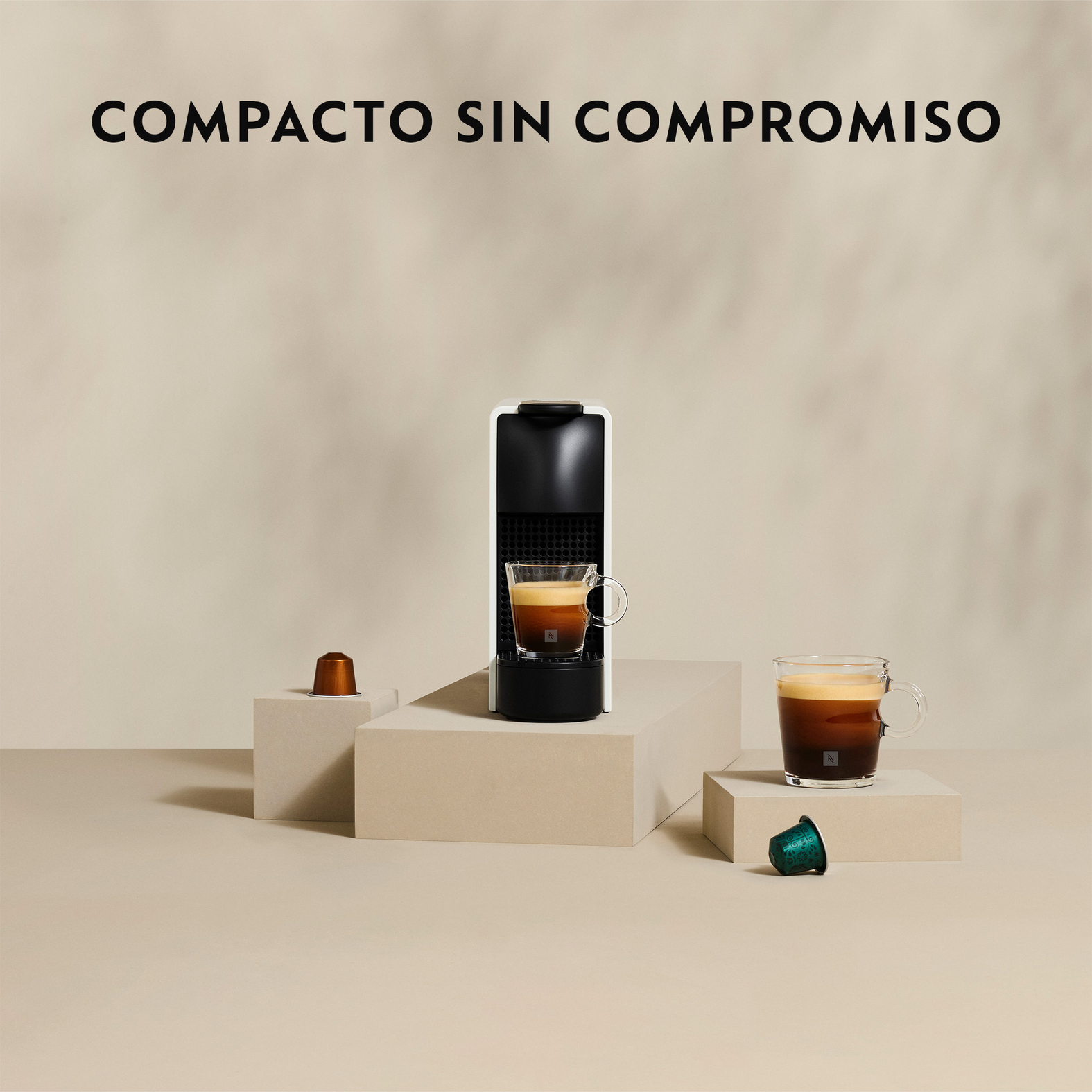 Una cafetera con dos tazas de café y cápsulas sobre pedestales beiges sobre fondo claro.