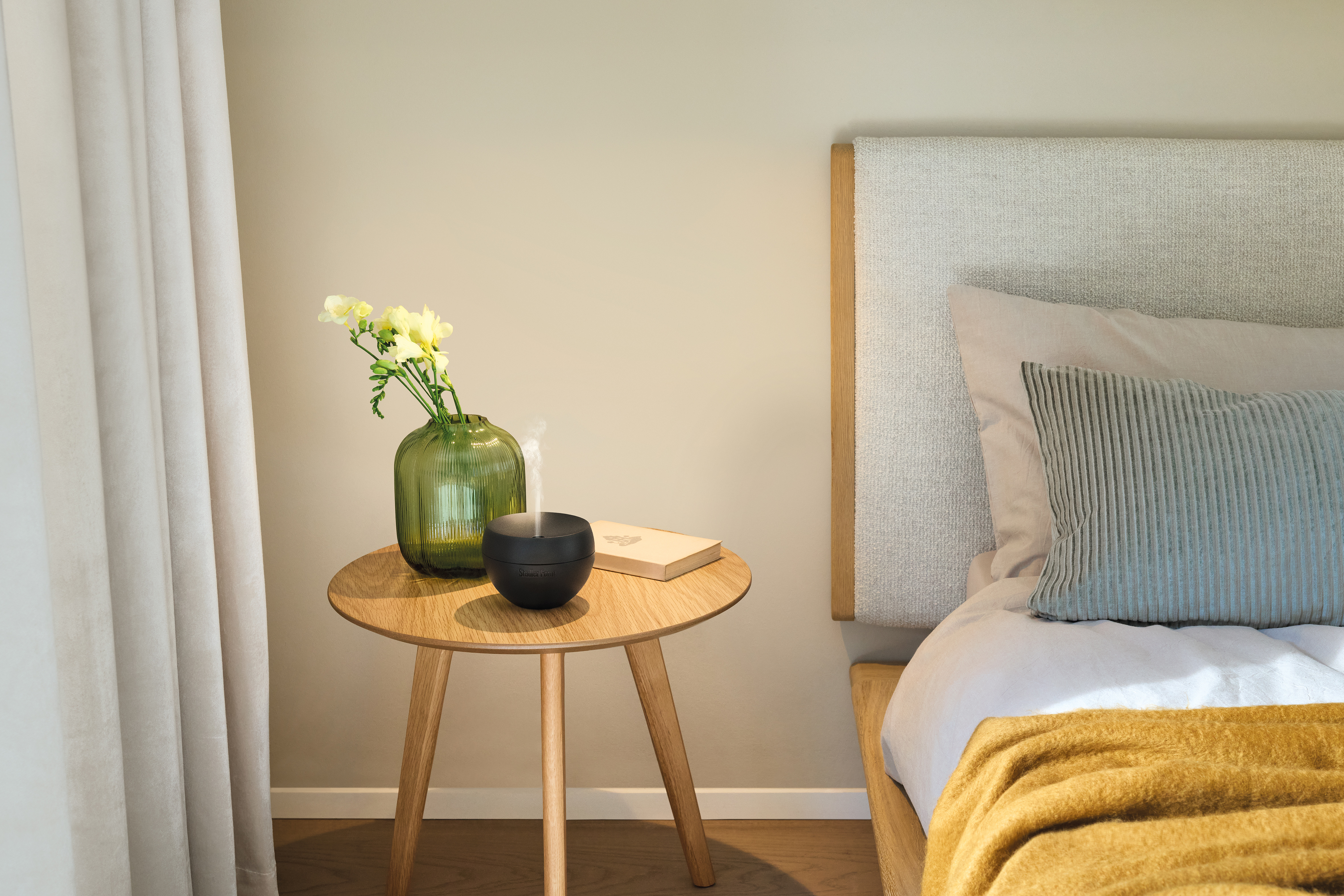 Ein Holztisch neben einem Bett mit einer grünen Vase, gelben Blumen, einem Buch und einem schwarzen Diffusor.