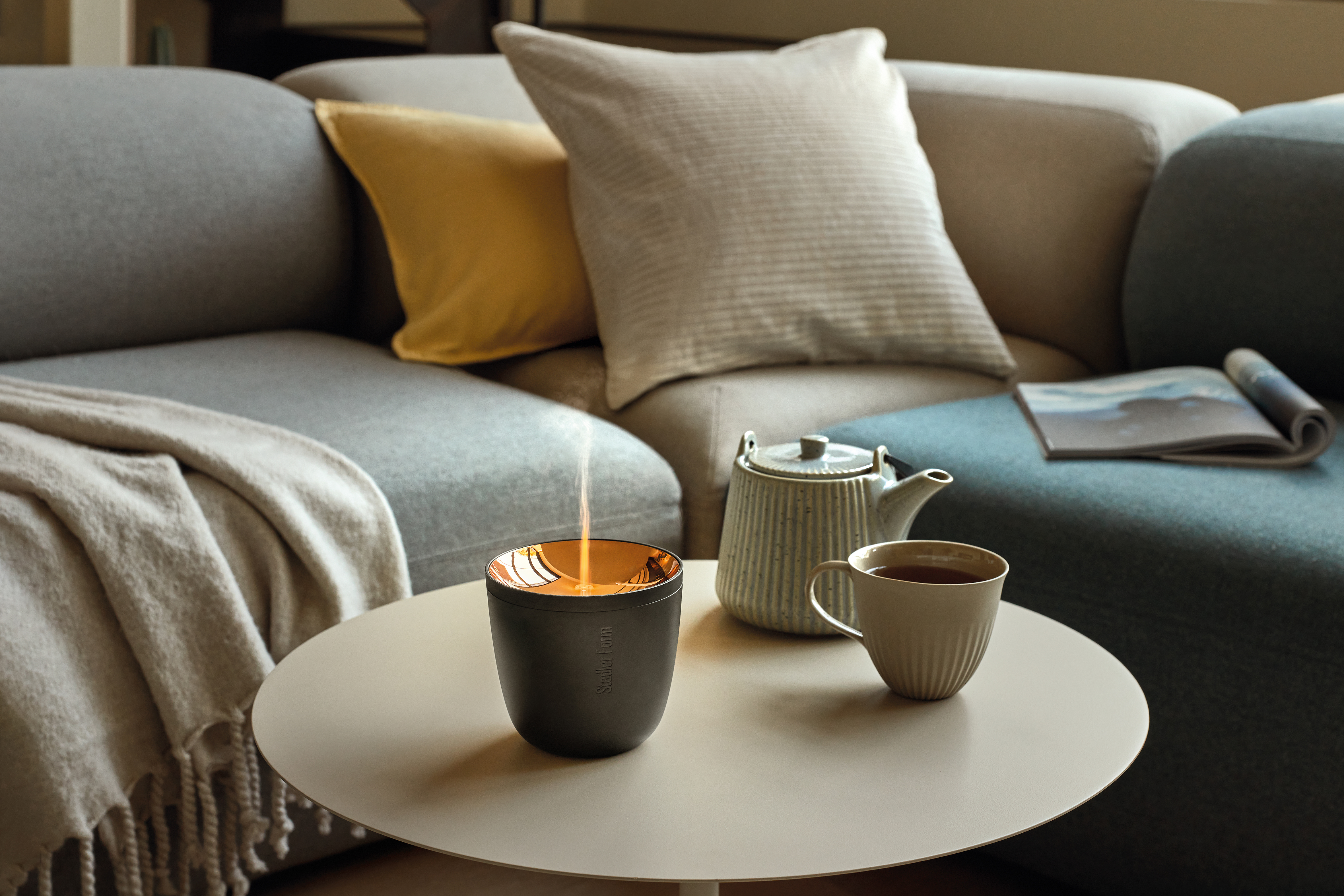 Runder Tisch mit Diffusor, Teekanne und Tasse vor einem grauen Sofa mit Kissen.