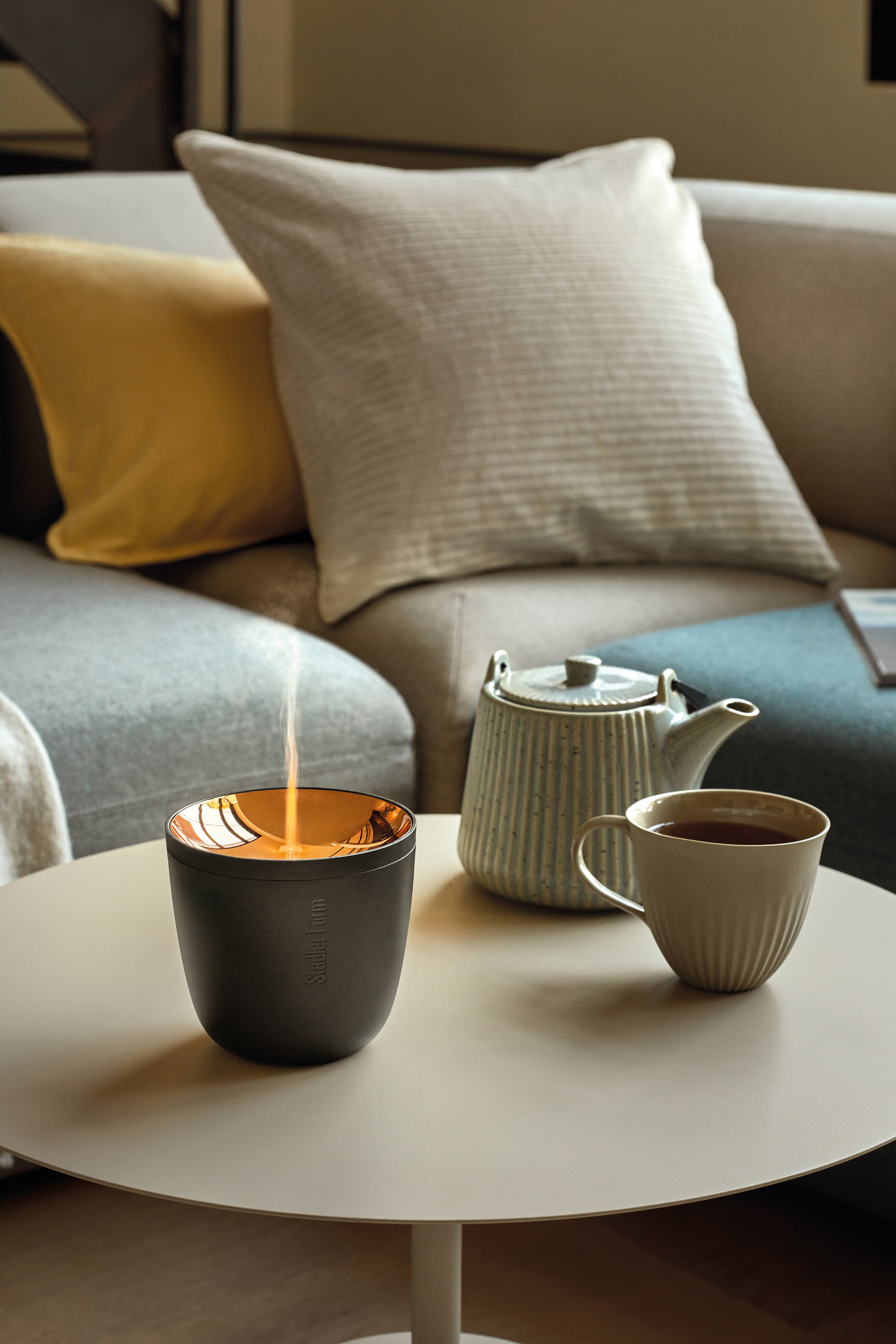 Ein Diffusor mit Flammen-Effekt, eine Teekanne und eine Tasse Tee stehen auf einem runden Tisch vor einem Sofa.