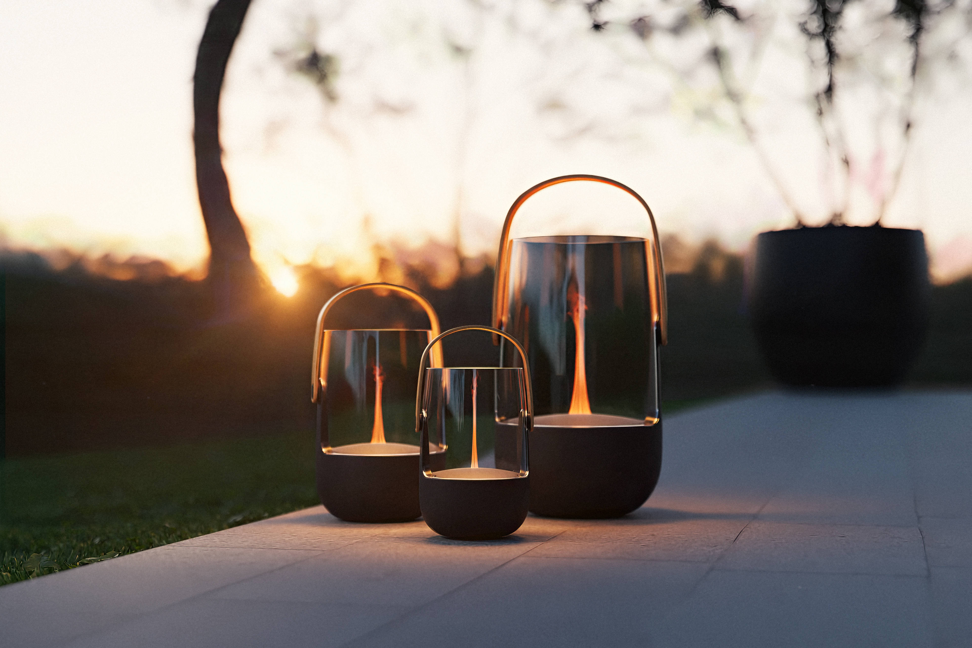 Drei moderne Glas-Laternen mit brennenden Flammen stehen im Freien bei Sonnenuntergang.