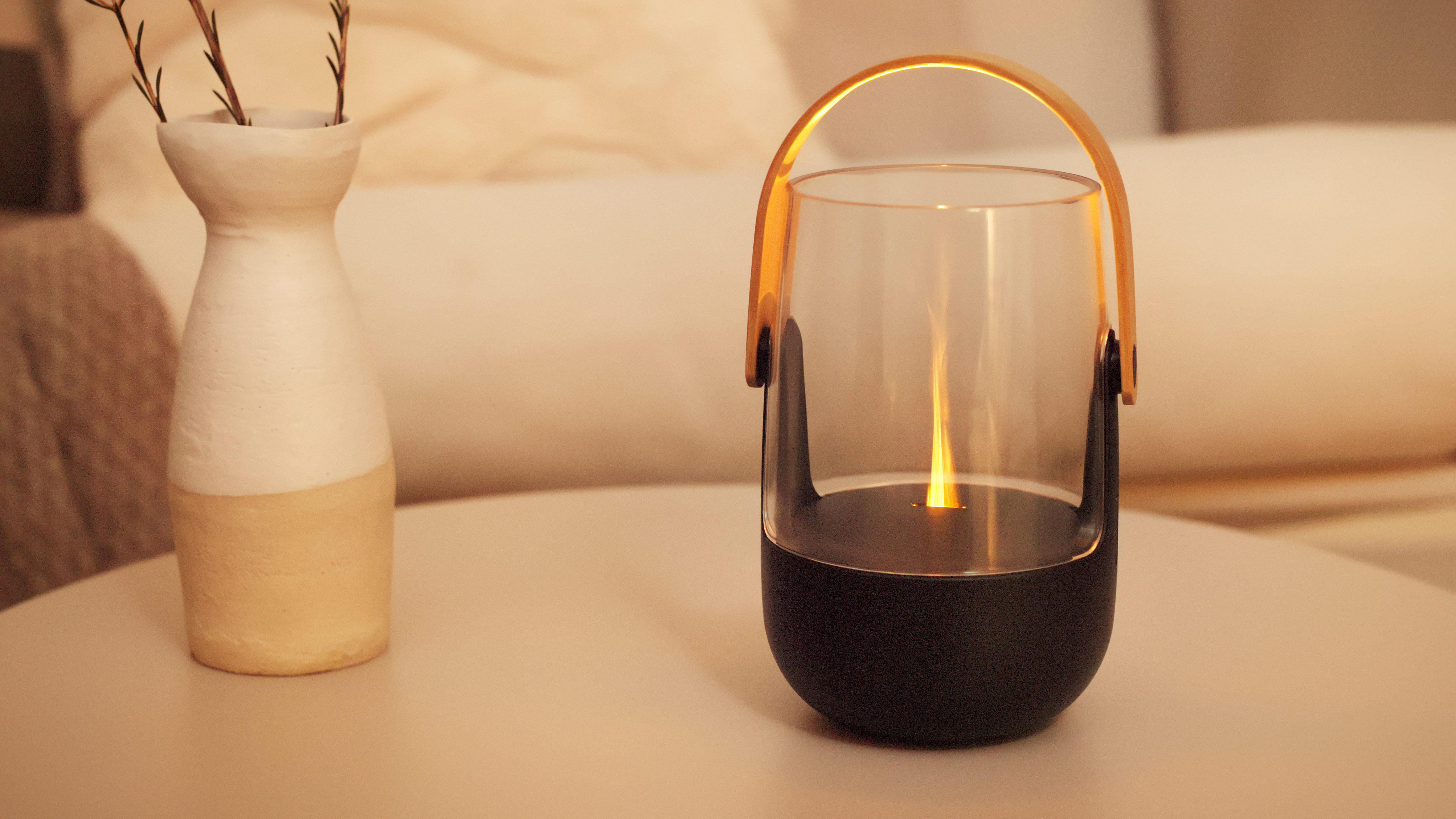 Eine moderne Glaslaterne mit schwarzem Sockel und goldenem Griff steht auf einem Tisch neben einer weißen Vase.
