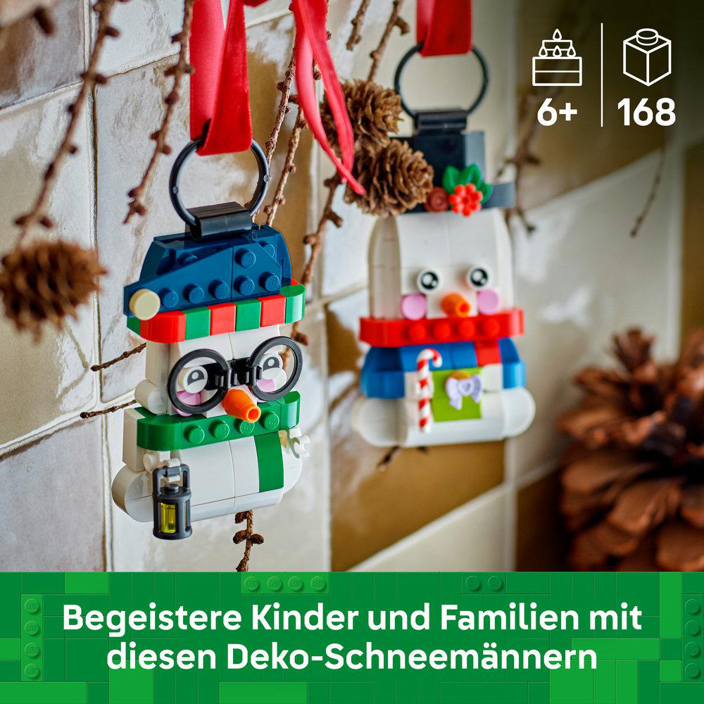 Zwei gebaute LEGO Schneemann-Ornamente hängen an roten Bändern, mit weihnachtlicher Deko im Hintergrund.