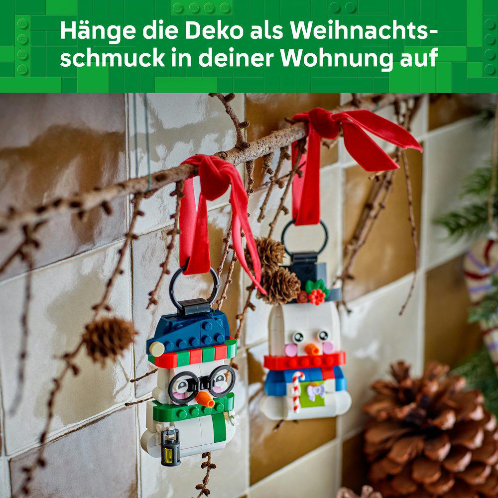Zwei LEGO Schneemann-Ornamente hängen an roten Schleifen an einem Ast vor einer gekachelten Wand.