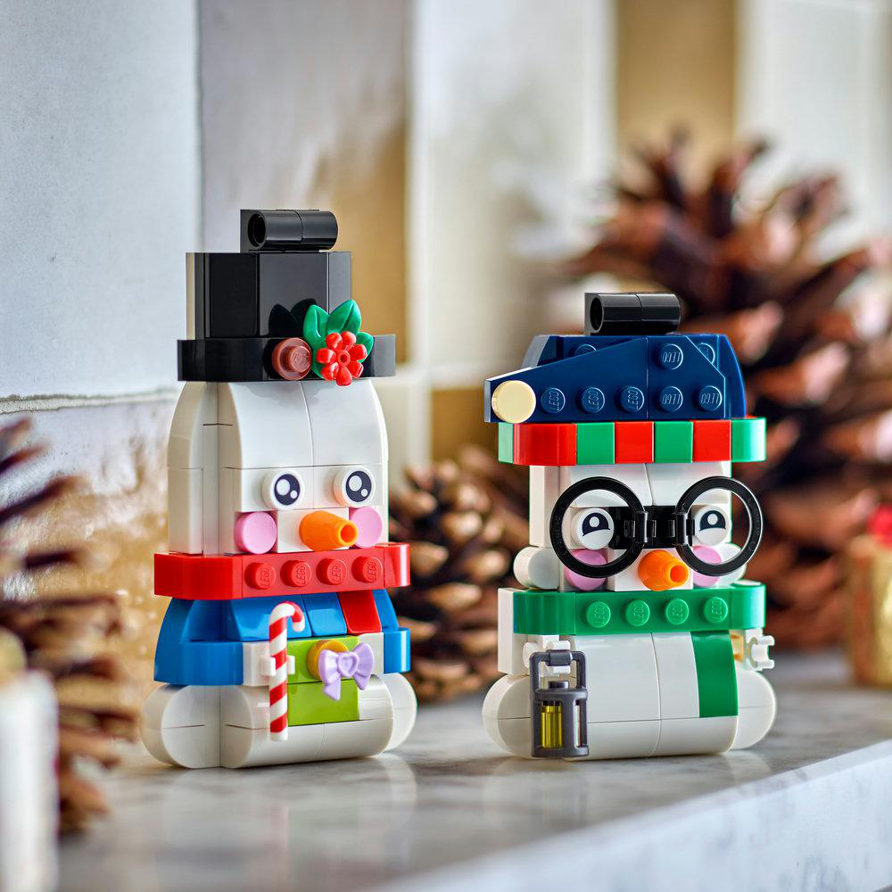 Zwei gebaute LEGO Figuren als Schneemänner mit Hüten und Schals vor Pinienzapfen.
