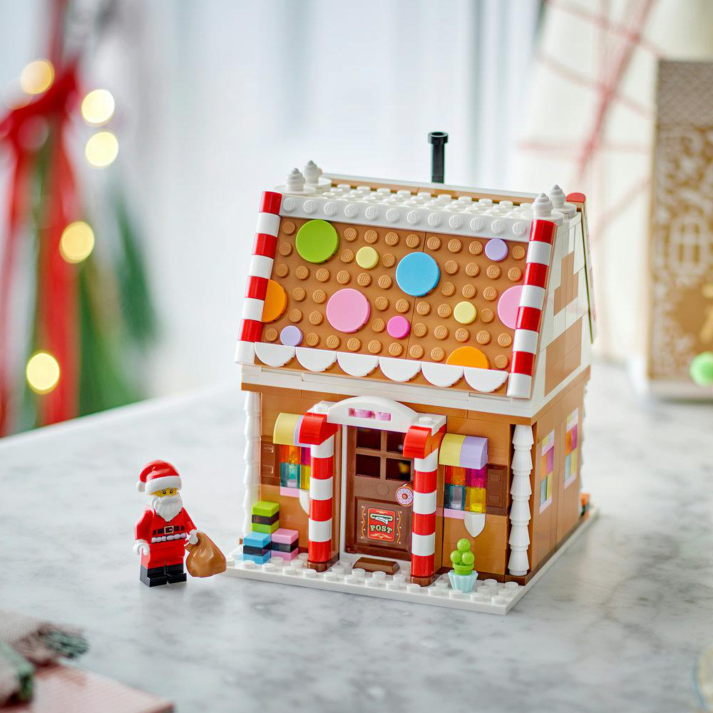 Lego-Lebkuchenhaus mit bunten Verzierungen, weißem Dach und einer Weihnachtsmann-Figur davor auf einem Tisch.