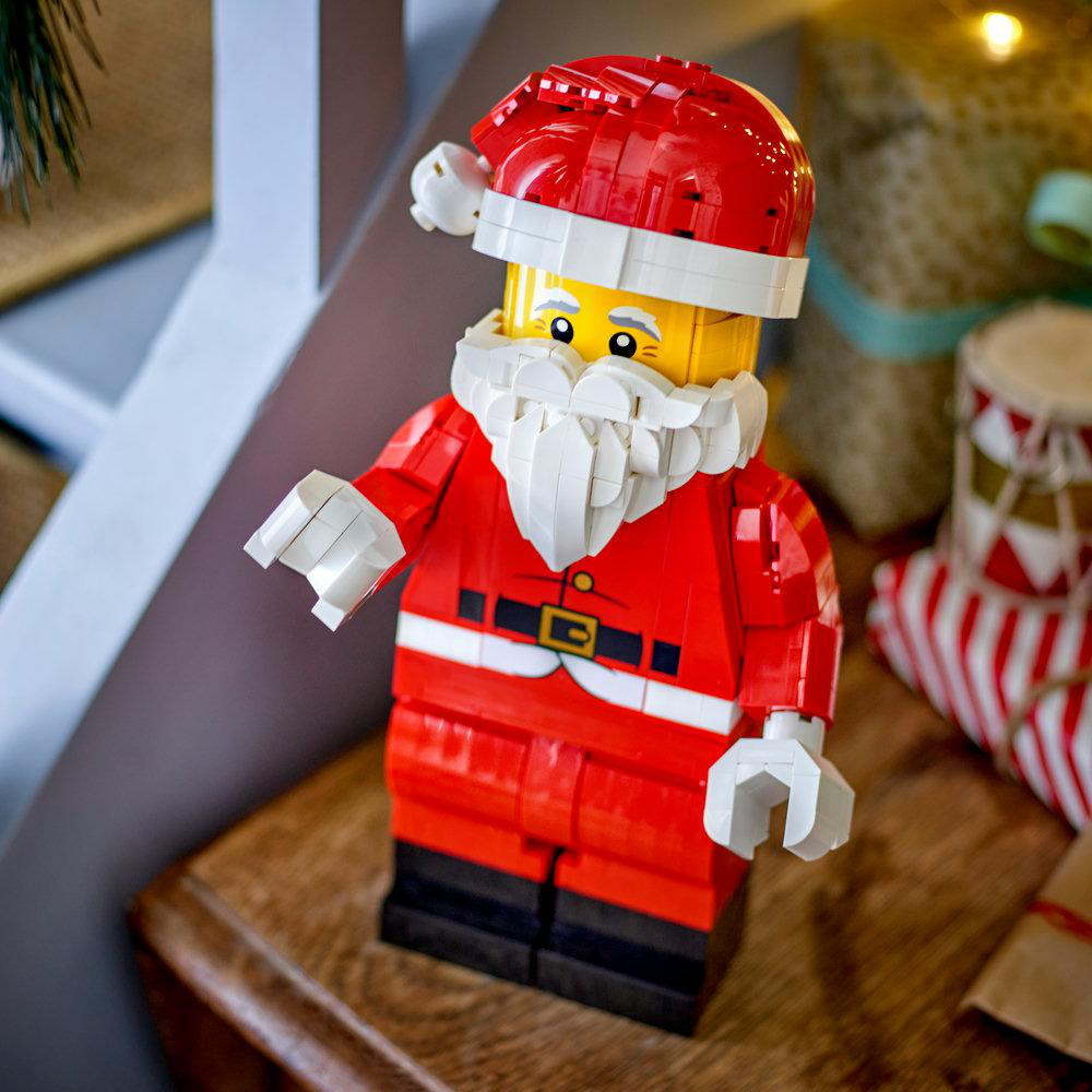 Eine Lego-Weihnachtsmannfigur steht auf einem Holzbrett vor verschwommenen Geschenken.