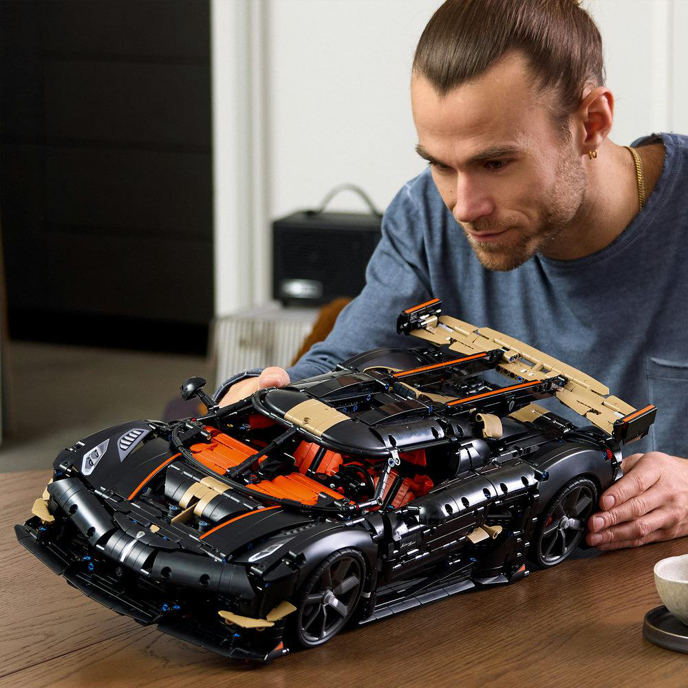 Ein Mann sitzt an einem Holztisch und baut ein großes, schwarzes Lego-Technic-Auto mit orangen Akzenten.