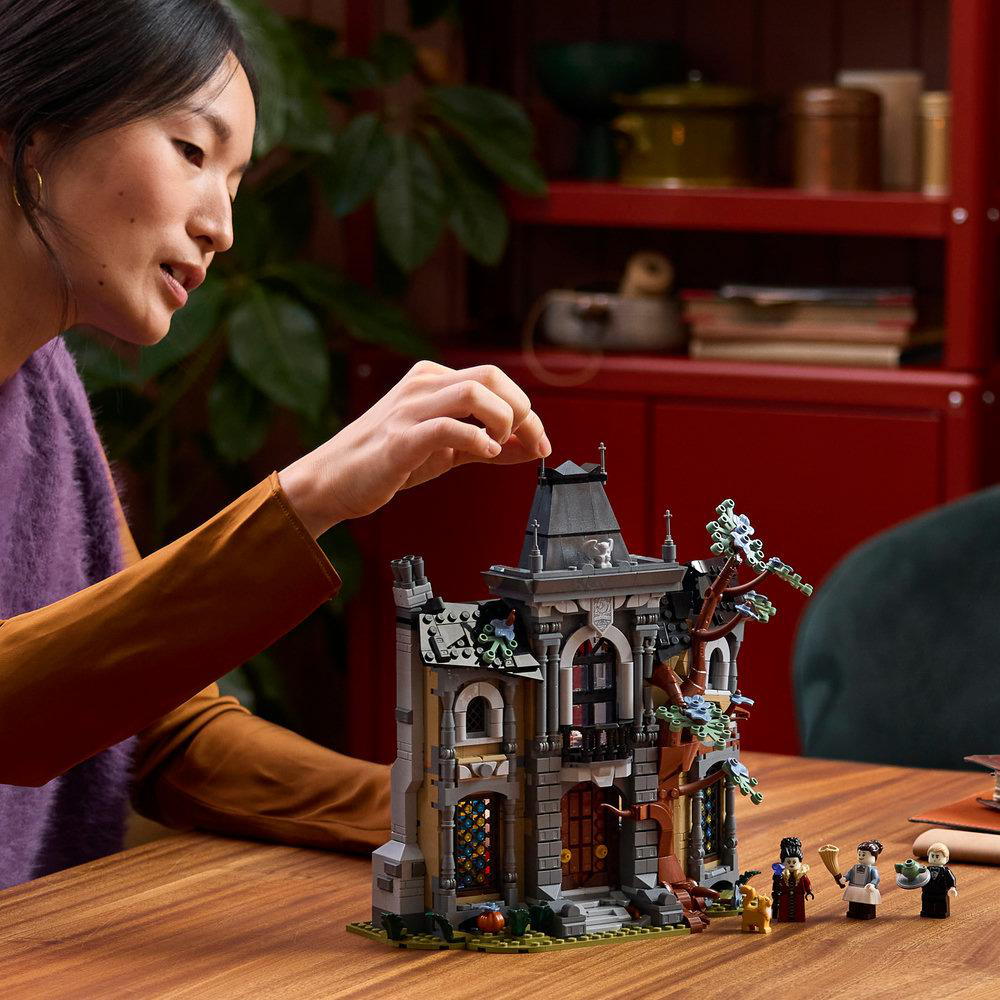 Eine Frau baut ein detailreiches Lego-Schloss auf einem Holztisch, im Hintergrund ist ein rotes Regal zu sehen.
