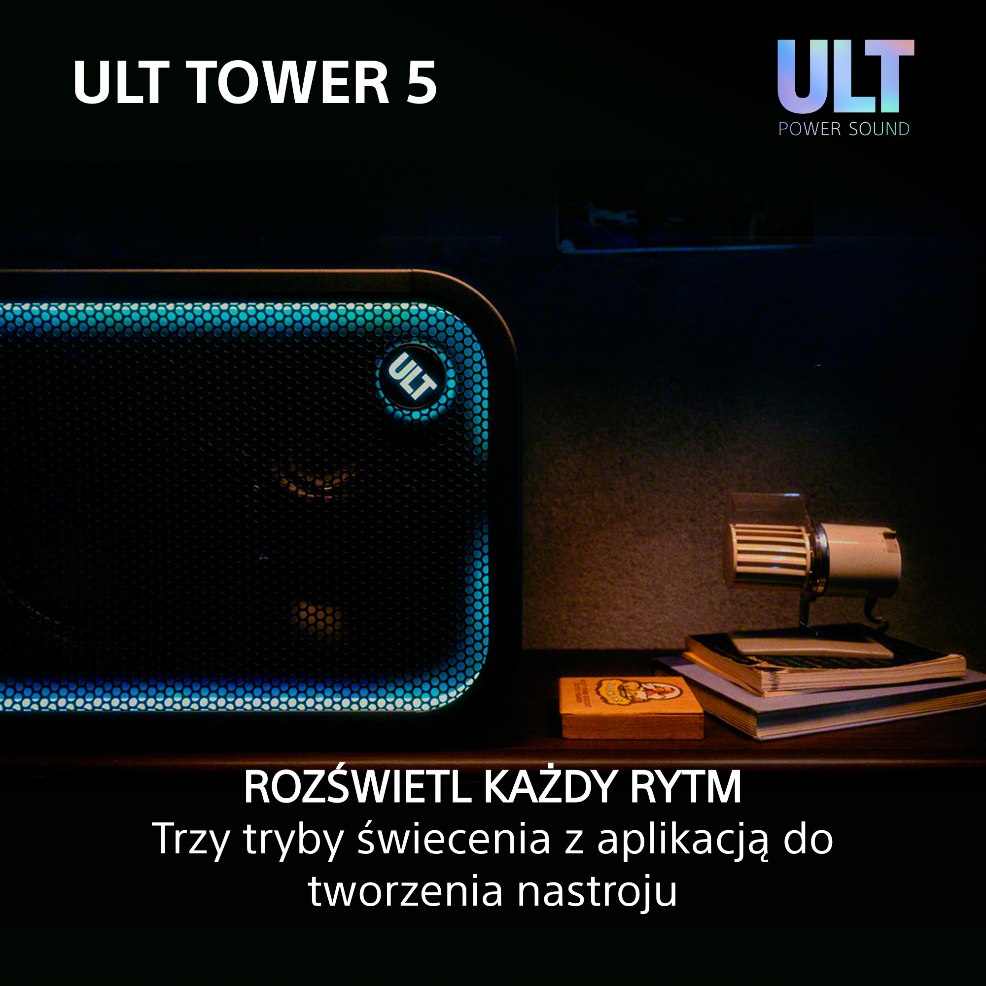 Głośnik ULT Tower 5 z niebieskim podświetleniem krawędzi stoi na drewnianym stole z książkami i lampką.